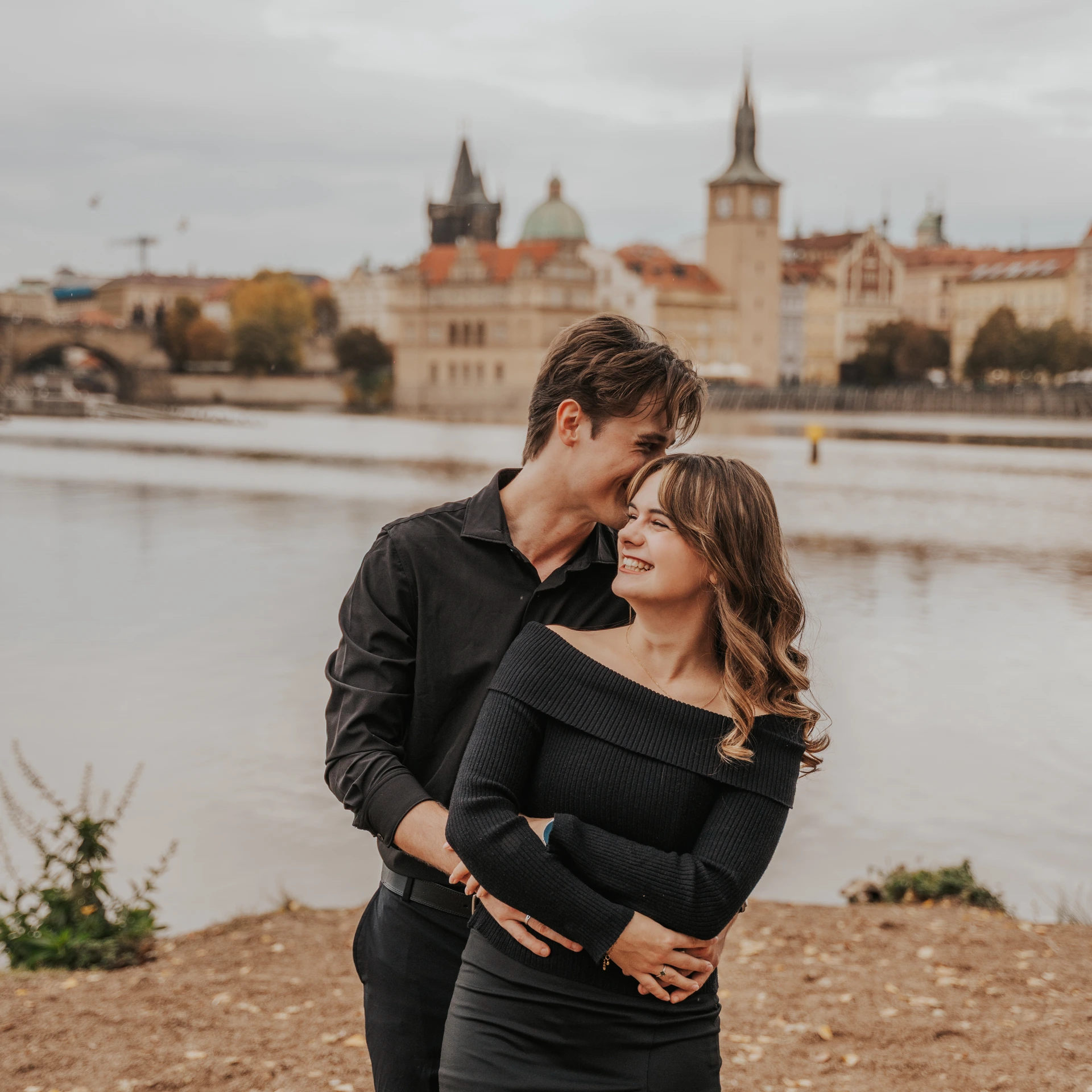 Couple in black clothing embracing beside the Vltava, with Prague’s towers and red rooftops in the background.