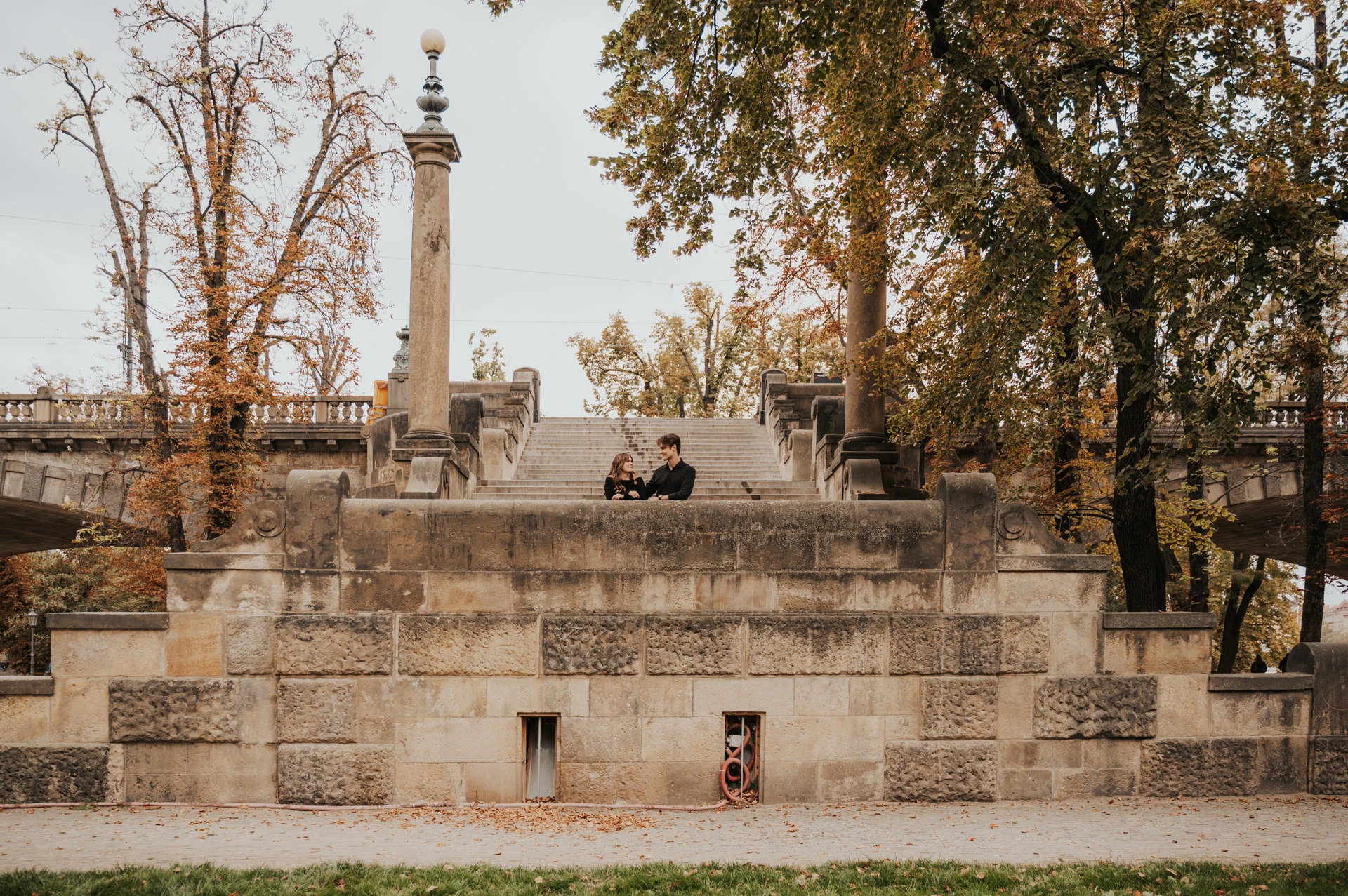 Couple looking at each other behind a stone balustrade in front of broad steps in Prague, with tall lamps and trees in autumn colours.