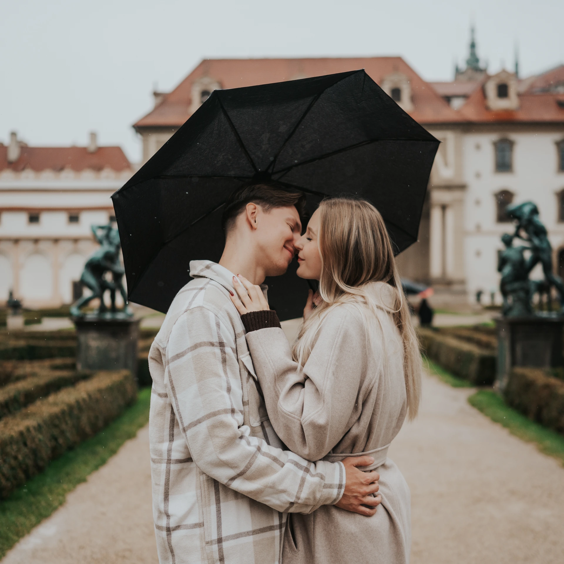 Couple standing close together under a black umbrella in Wallenstein Garden in Prague, between hedges and bronze statues.