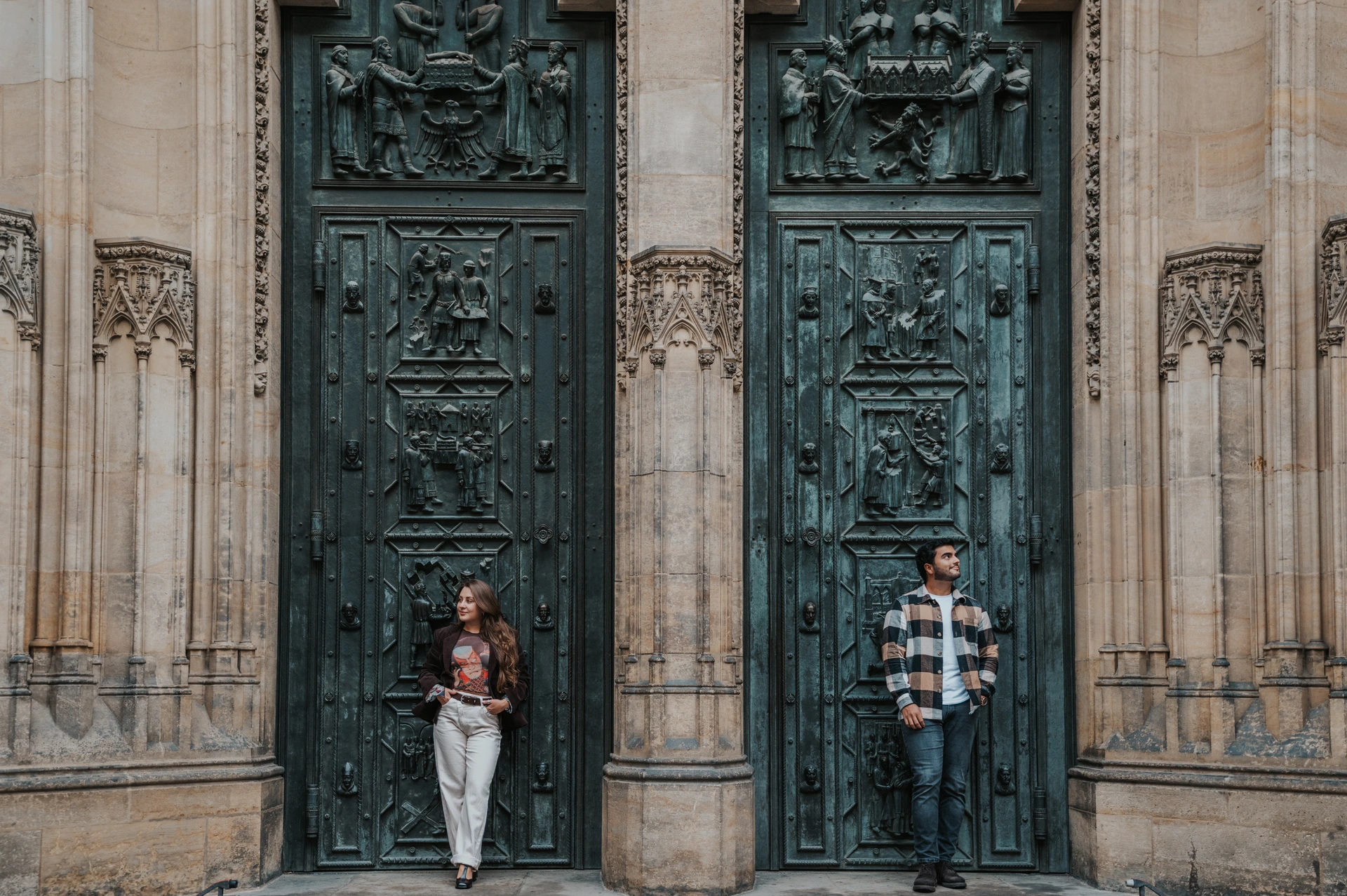 Couple standing on either side of a stone column in front of tall bronze doors with relief panels and Gothic carvings in Prague.