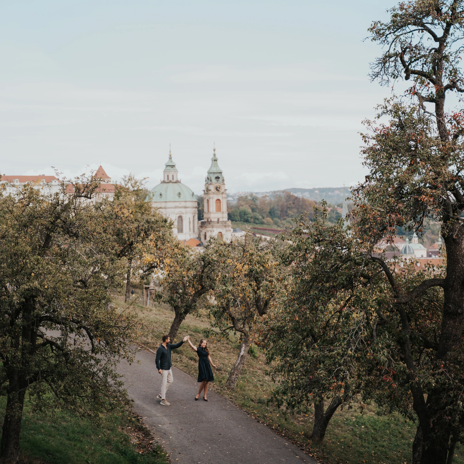 Couple dancing on a path between fruit trees, with a church dome, tower and Prague’s red rooftops in the background.