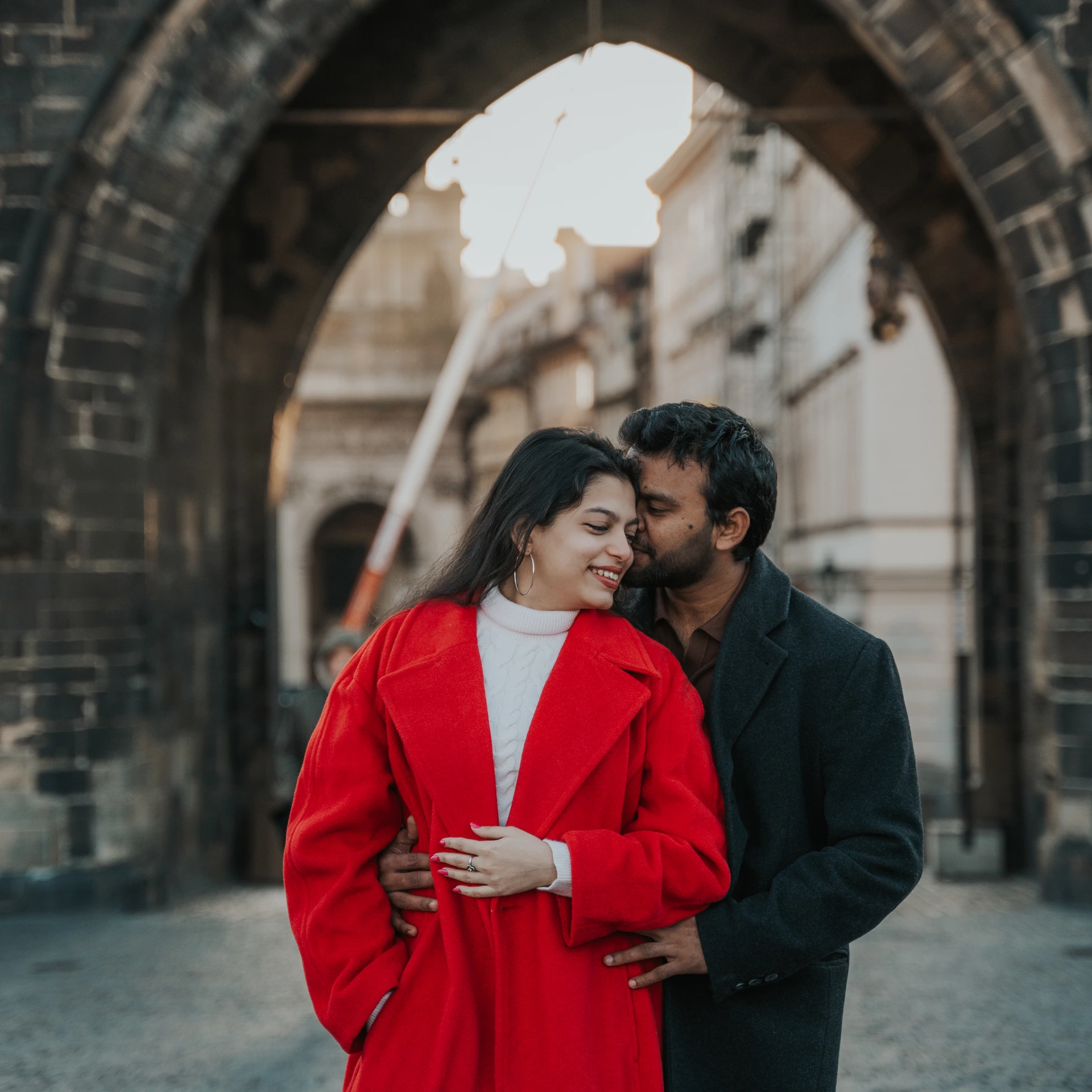 Couple embracing beneath a Gothic stone arch in Prague; she is wearing a red coat.