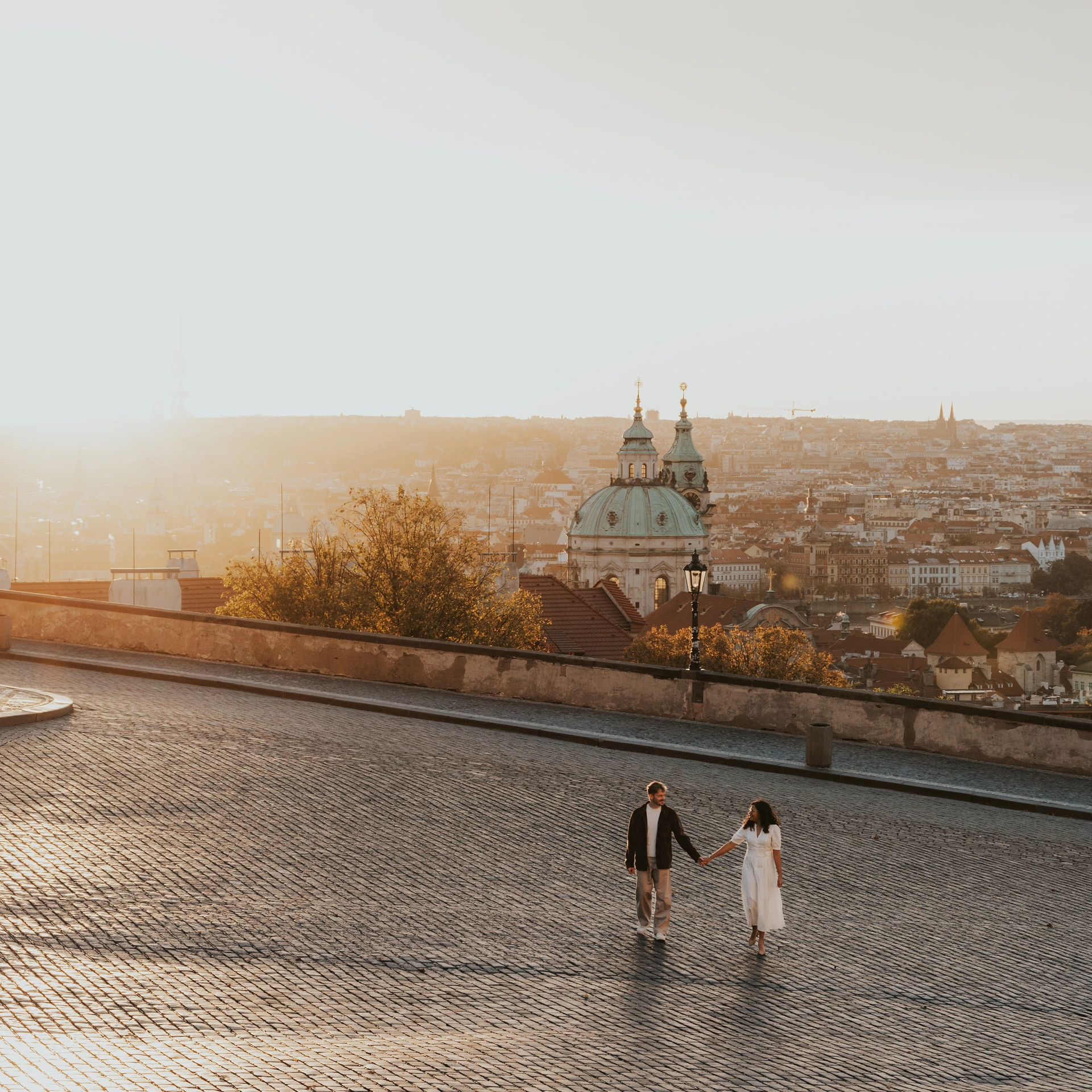 Couple walking hand in hand across a broad cobbled square overlooking Prague, a green church dome and the skyline in warm sunlight.