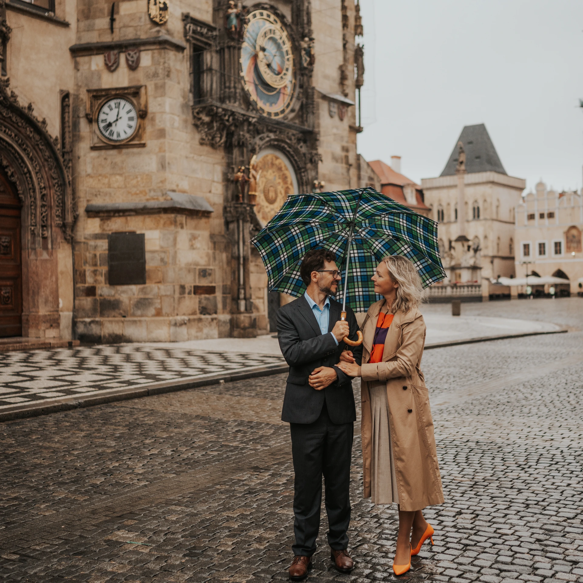Couple sharing a green-and-blue checked umbrella beside the Astronomical Clock in Prague Old Town Square.