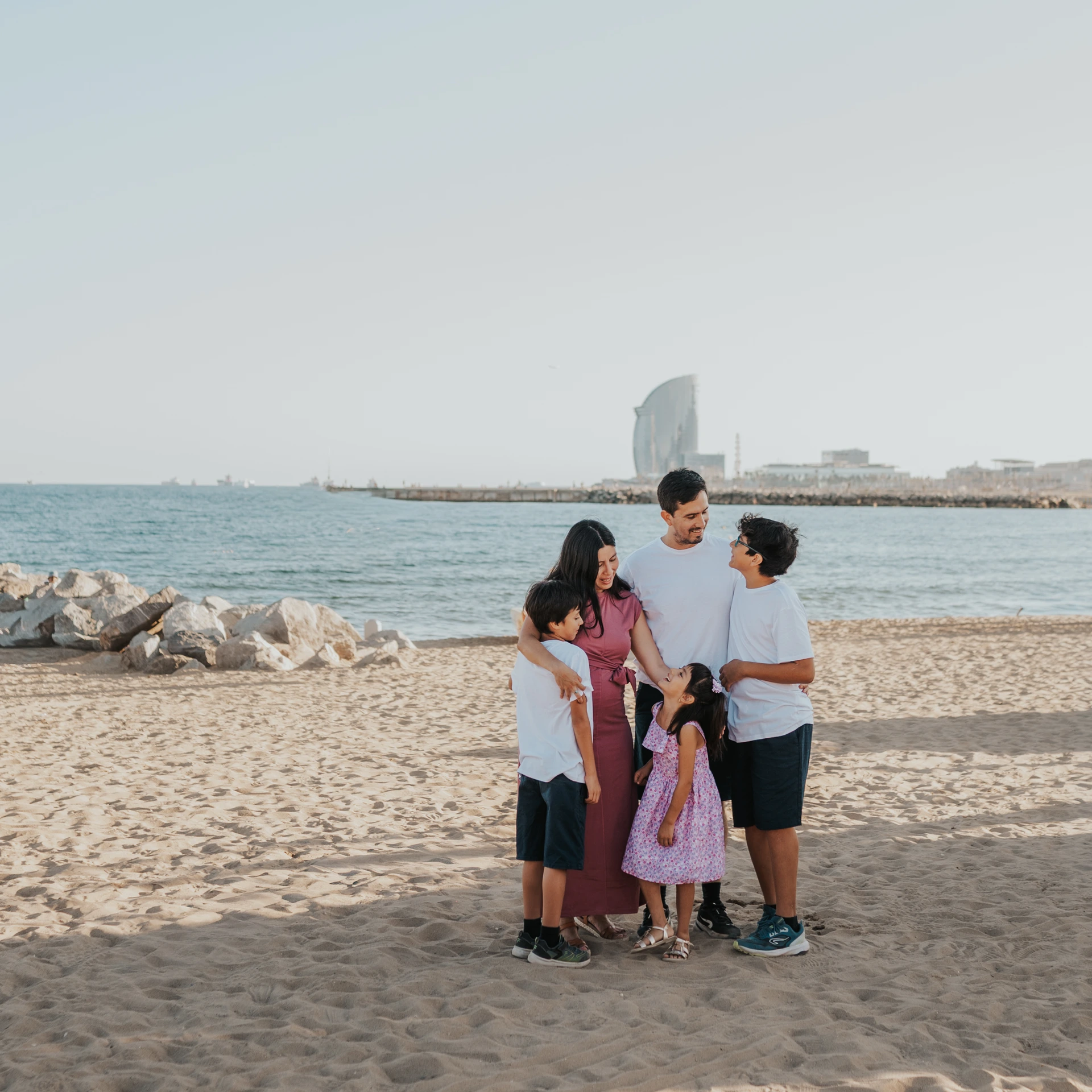 Rodzina stoi blisko siebie na plaży Barceloneta, a po drugiej stronie wody widać hotel W Barcelona podczas rodzinnej sesji zdjęciowej w Barcelonie.
