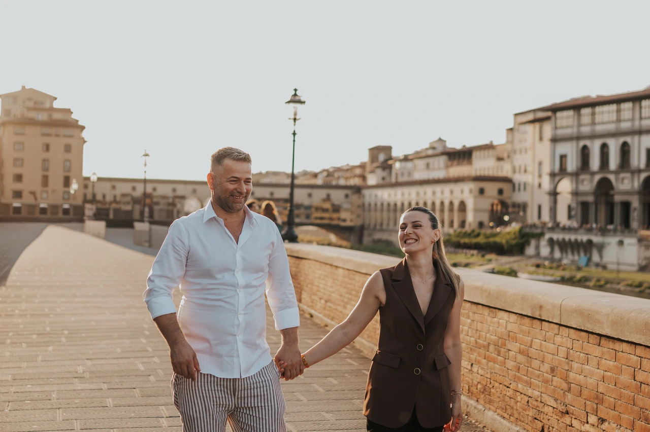Una coppia cammina mano nella mano accanto a un parapetto in mattoni lungo l’Arno durante un servizio fotografico a Firenze.