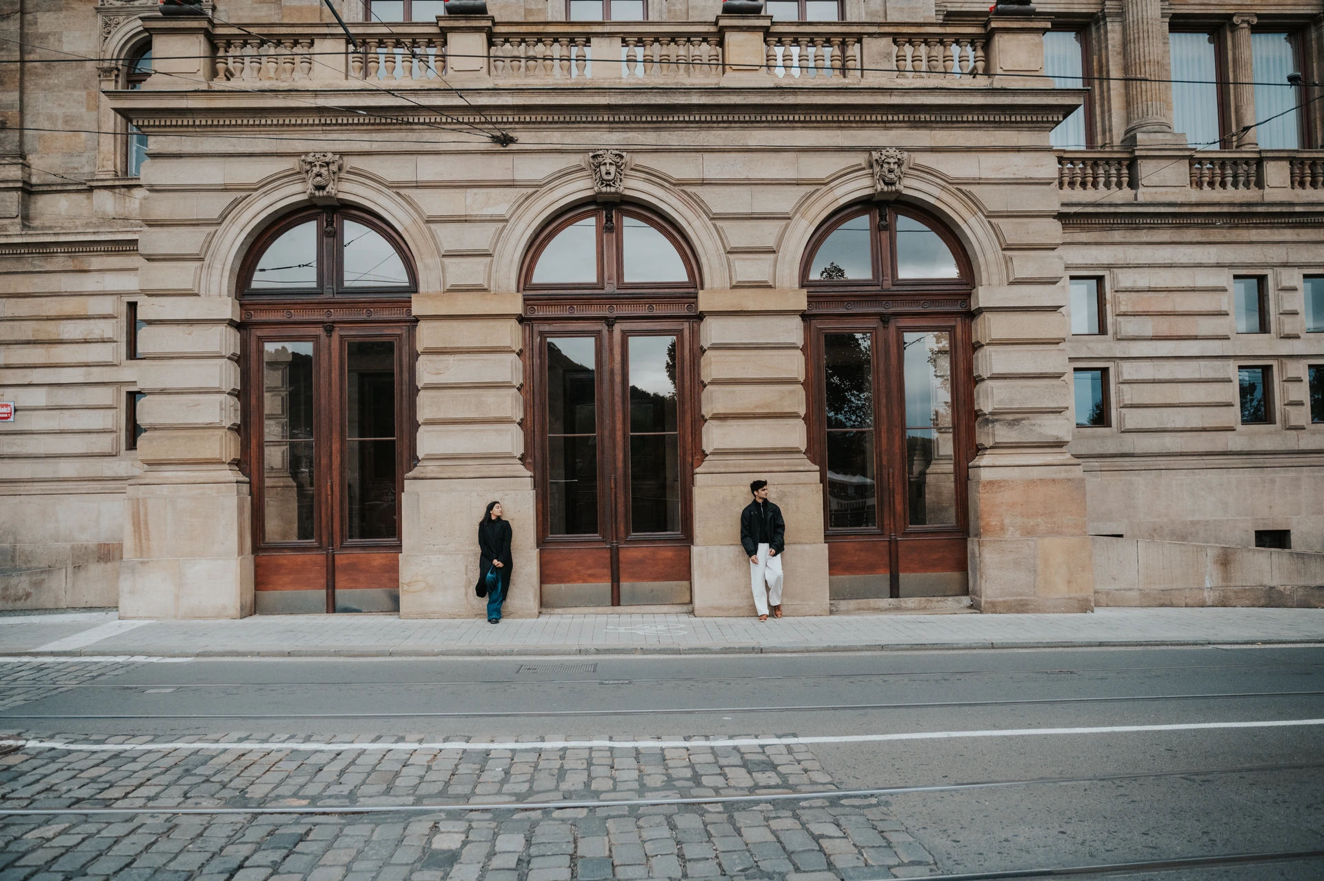 Couple posing a little apart in front of tall arched wooden doors in a sandstone facade in Prague, with tram tracks in the foreground.