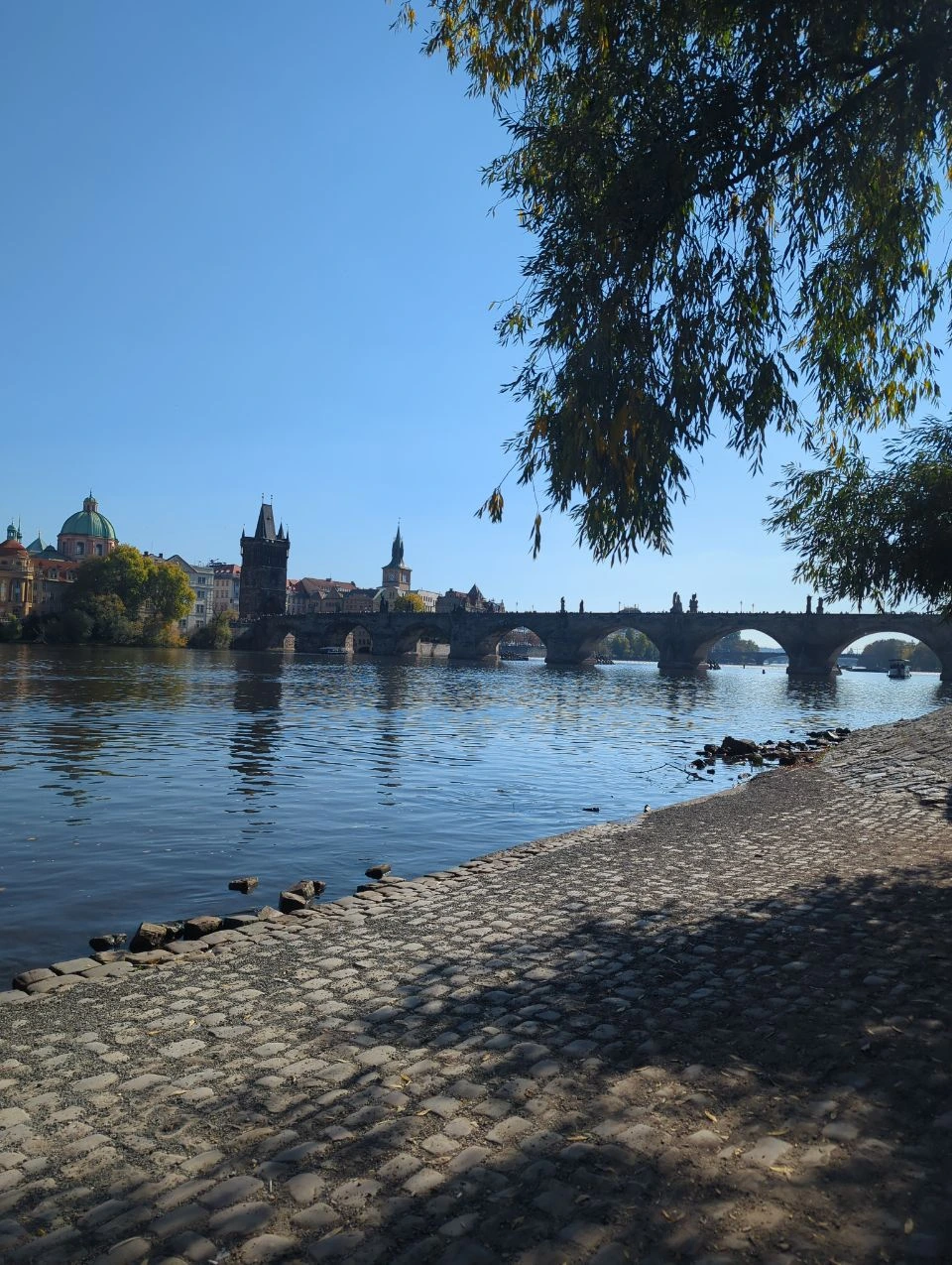 Puente de Carlos desde una orilla adoquinada y tranquila del Moldava en Praga