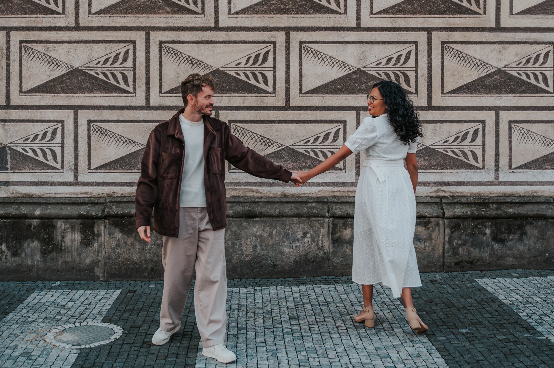 Couple holding hands on a cobbled street in front of a facade with geometric patterns in Prague; she wears a white dress and he wears a brown jacket.