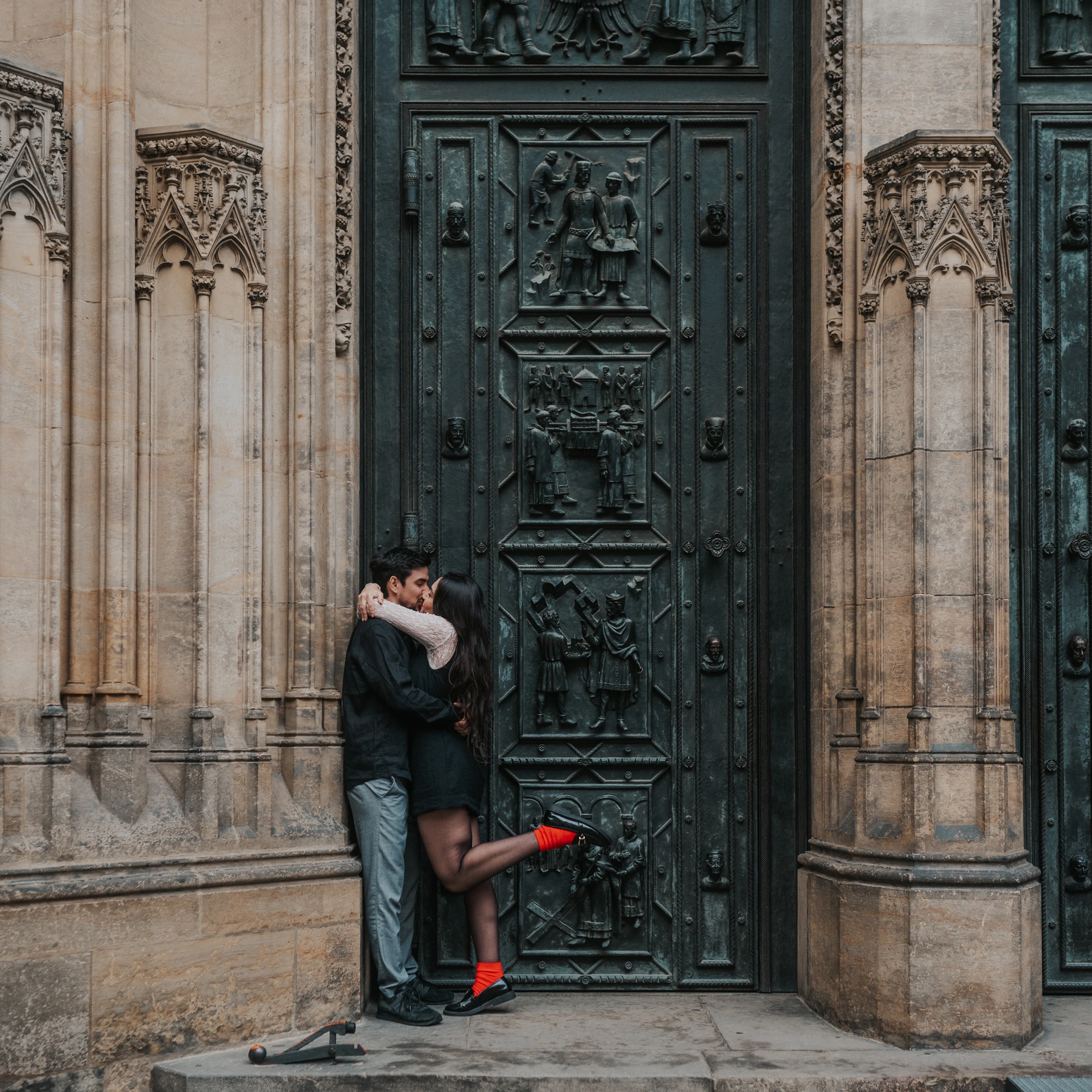 Couple kissing in front of a tall bronze door with relief panels and Gothic columns at Prague Castle.