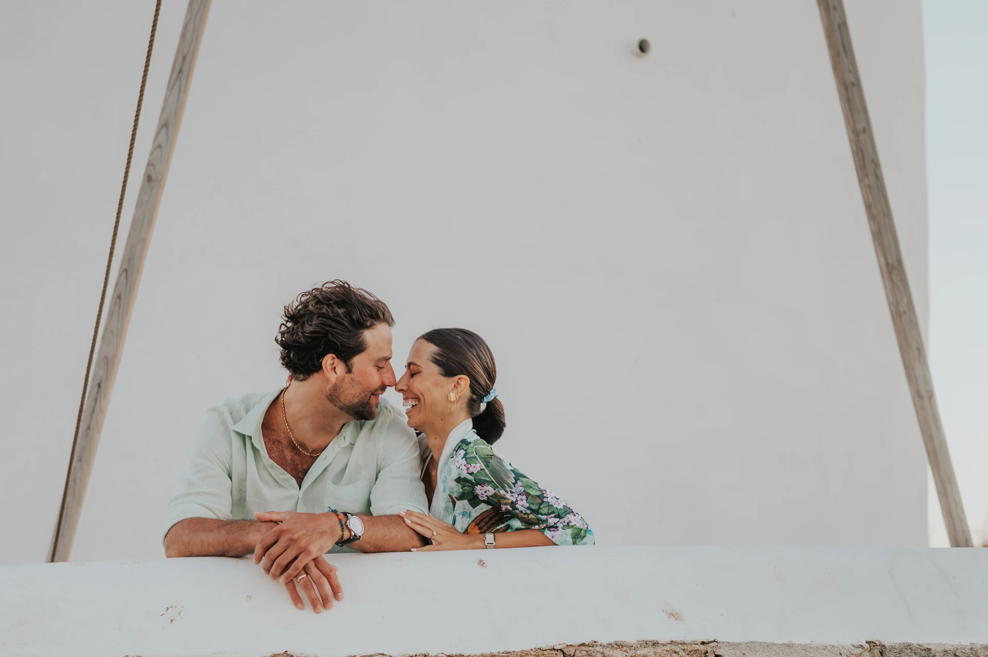 A smiling couple leans close across a white wall during a Mykonos couple photoshoot at the windmills.