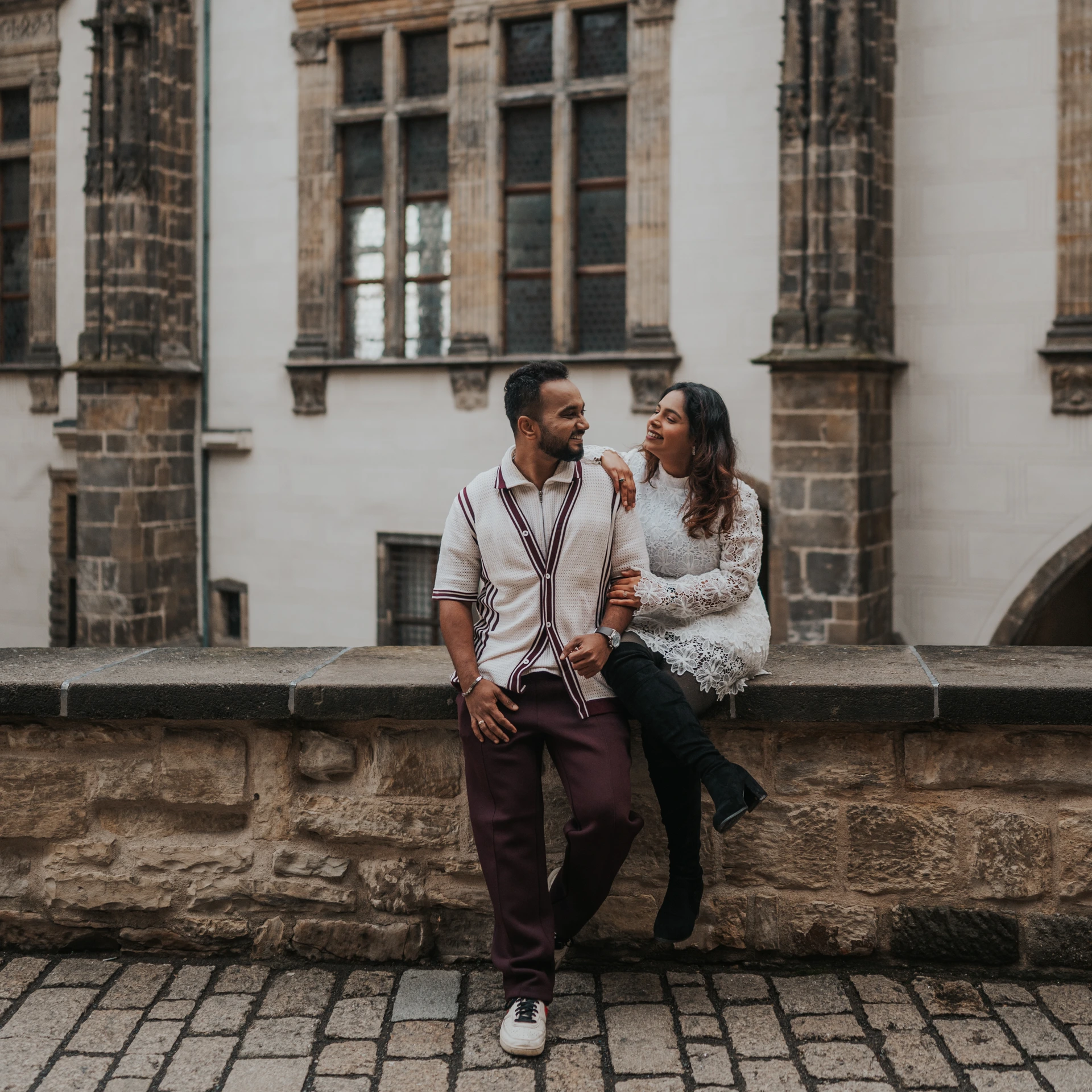 Couple smiling at each other beside a low stone wall in Prague, in front of a historic facade with tall windows.