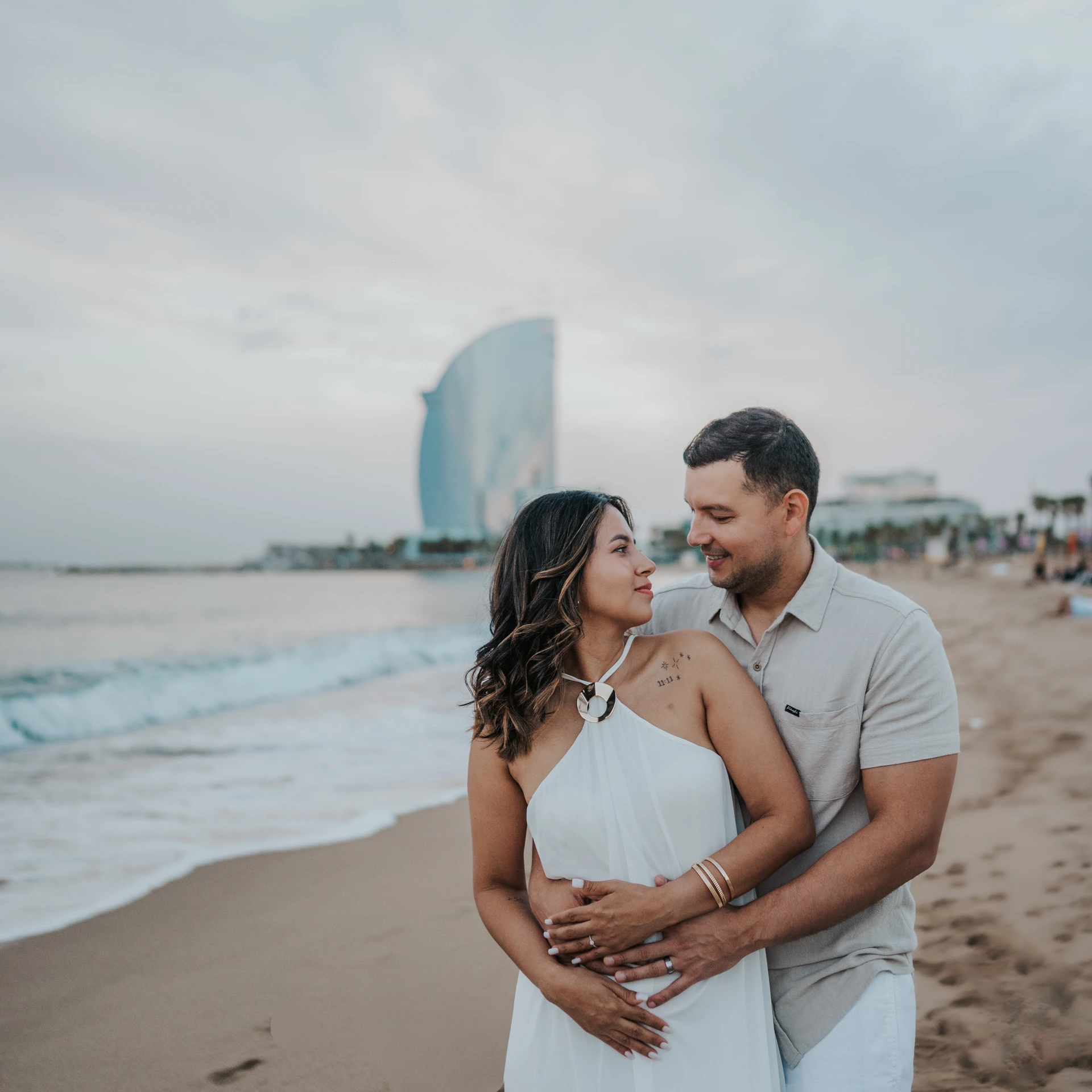 Przyszli rodzice obejmują się i trzymają ciążowy brzuch na plaży Barceloneta z hotelem W Barcelona w tle podczas sesji ciążowej w Barcelonie.