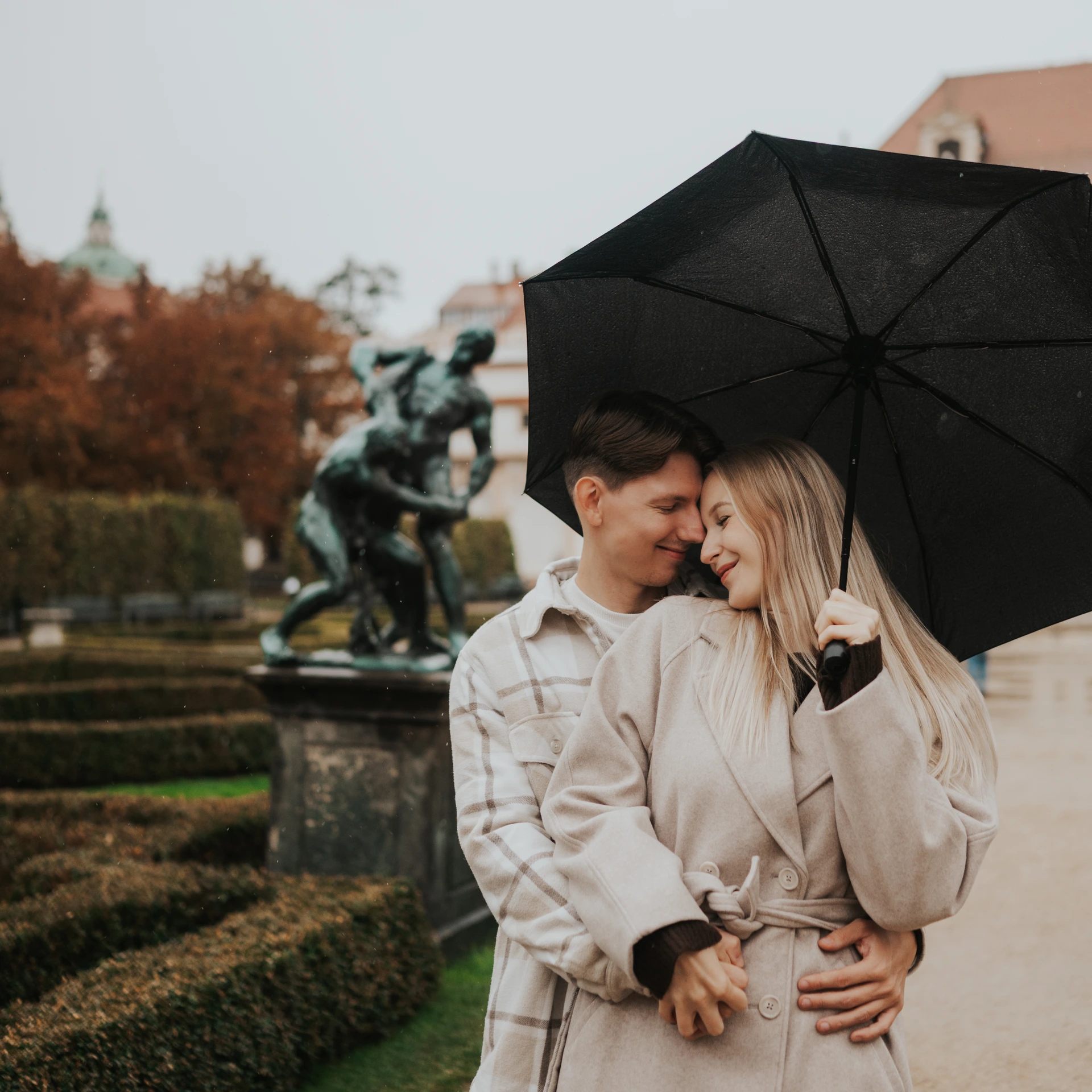 Couple standing under a black umbrella in Wallenstein Garden in Prague, with hedges and a bronze statue behind them.