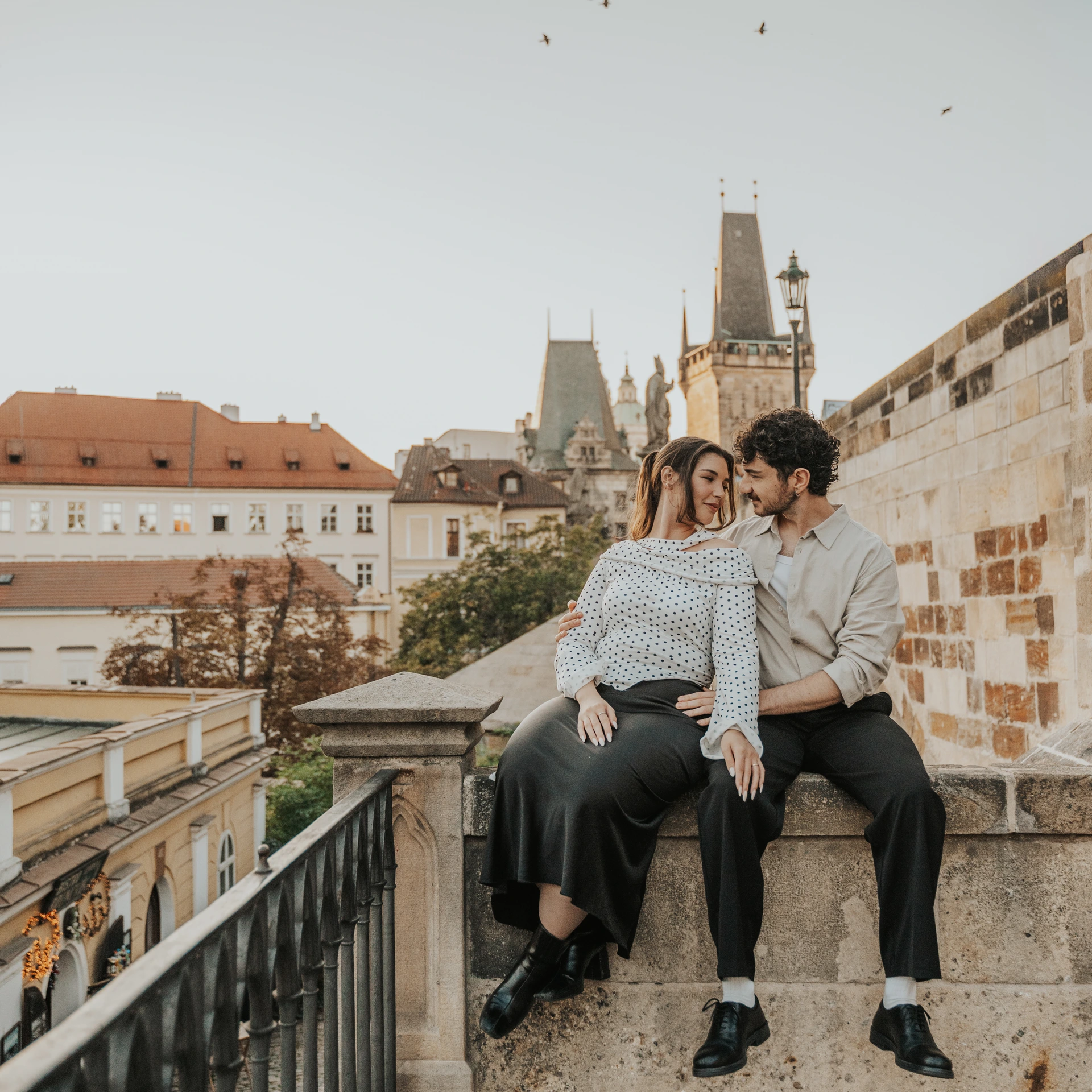 Couple sitting close together on a stone wall beside Charles Bridge in Prague, with the bridge towers behind them.
