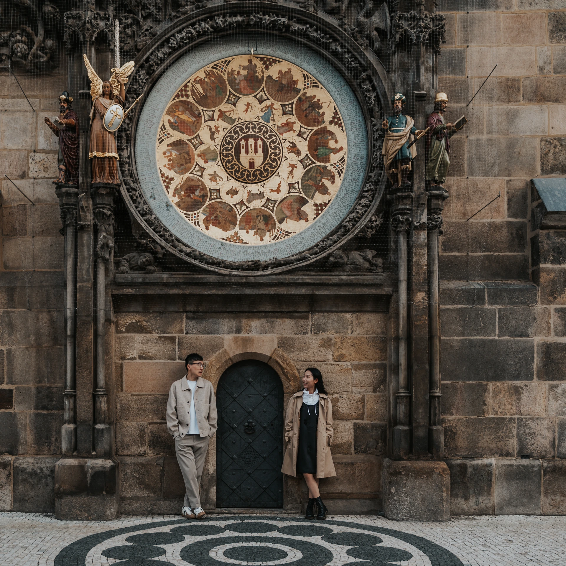 Couple in beige coats standing beneath the painted calendar dial of the Astronomical Clock in Prague Old Town Square.