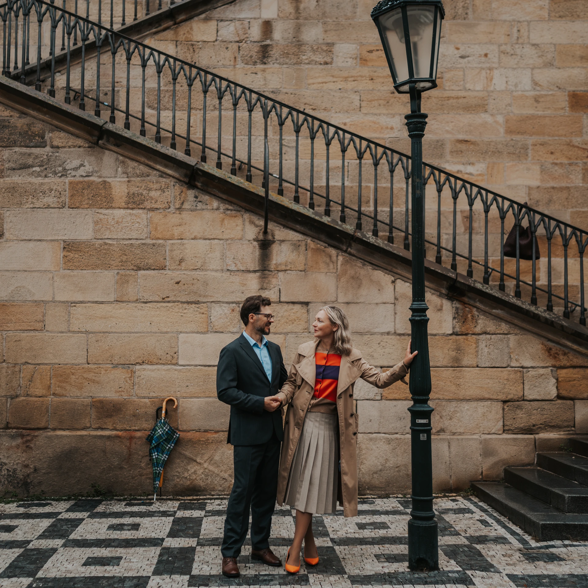 Couple holding hands beside a lamppost and stone steps in Prague; a checked umbrella rests against the wall.