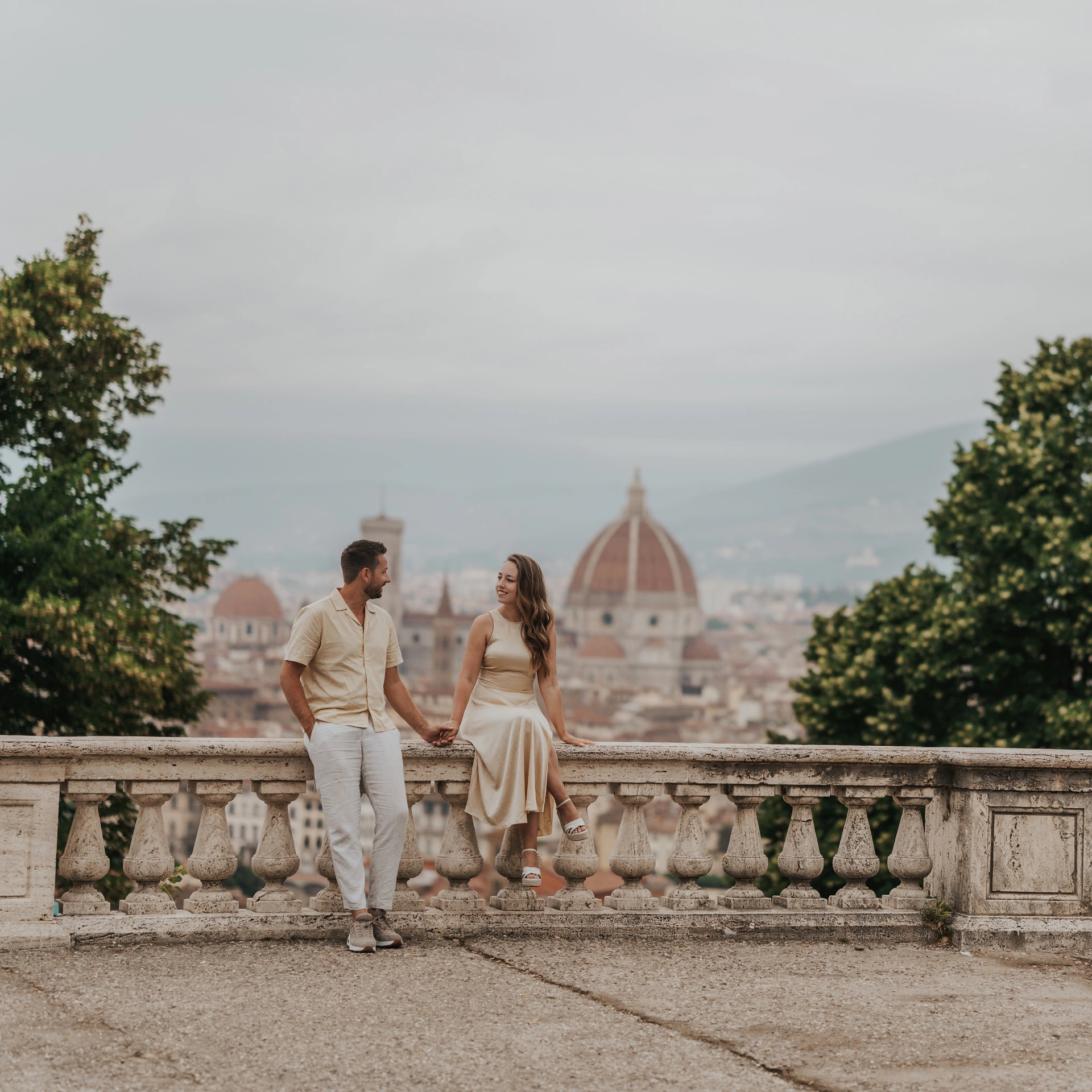 Una pareja se toma de la mano junto a una balaustrada de piedra, con el Duomo al fondo, durante una sesión de fotos en Florencia.