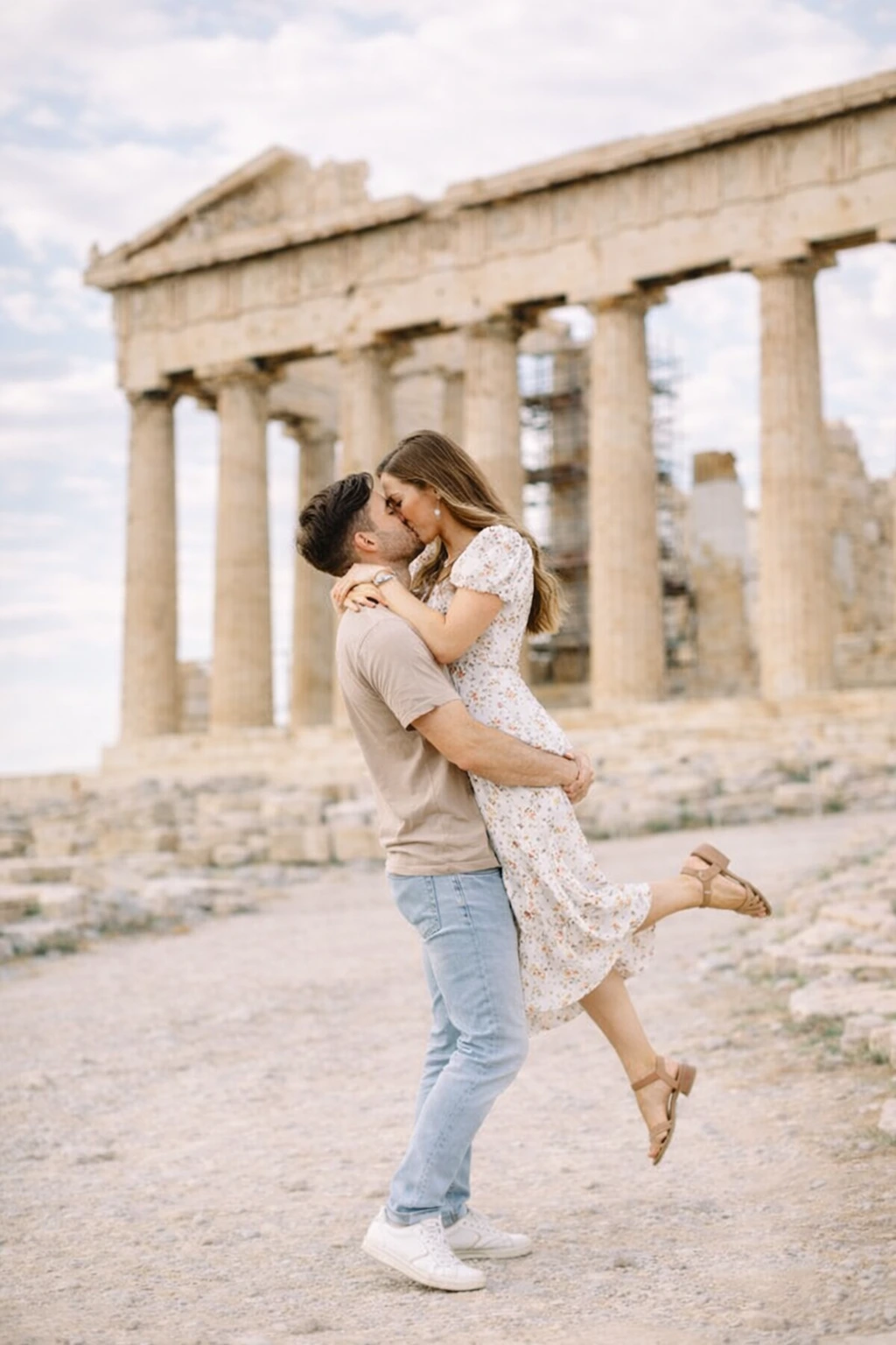 Two adults kiss as one lifts the other in a portrait with the Parthenon clearly visible behind them.
