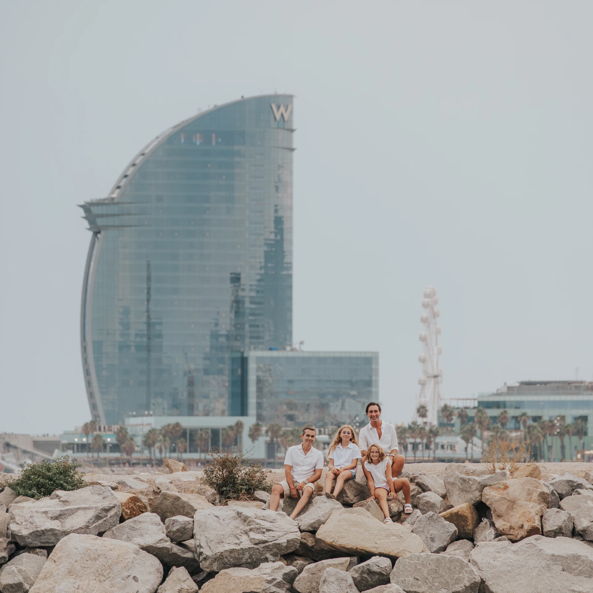 Een gezin zit samen op rotsen aan zee met het zeilvormige W Barcelona hotel op de achtergrond tijdens een gezinsfotoshoot in Barcelona.