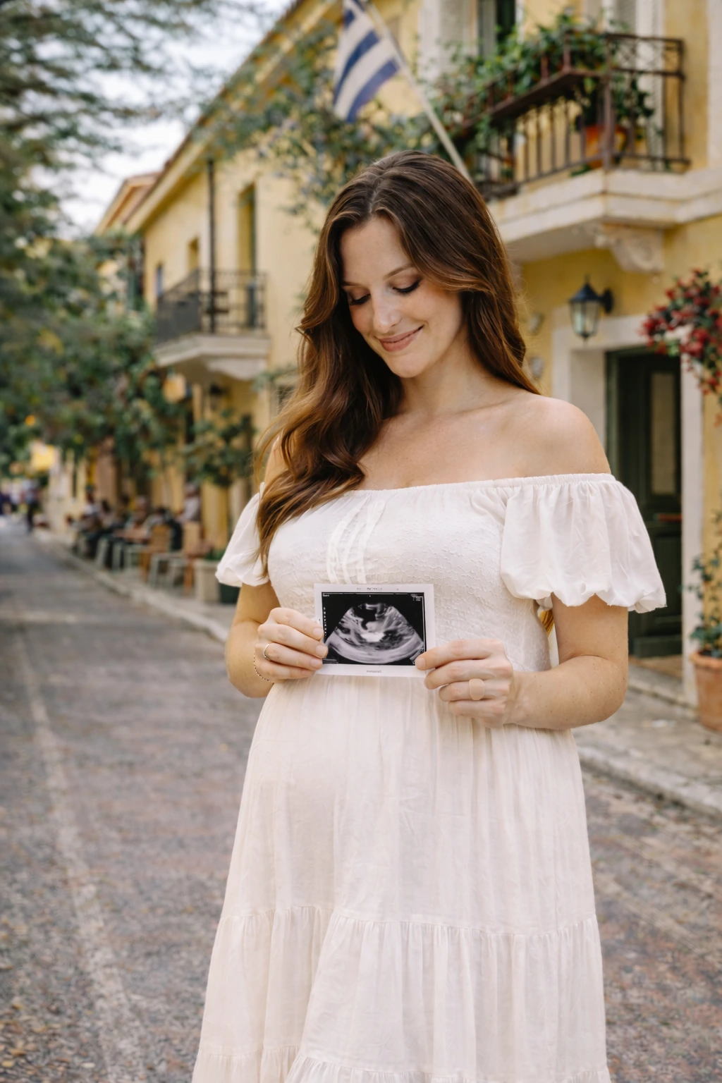 A woman in a flowing white off-shoulder dress holds an ultrasound image on a colourful Plaka street during an Athens photoshoot.