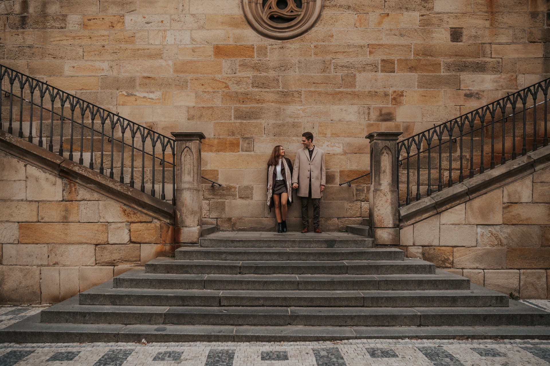 Couple holding hands on broad stone steps in Prague, between two symmetrical staircases with black railings in front of a sandstone facade.