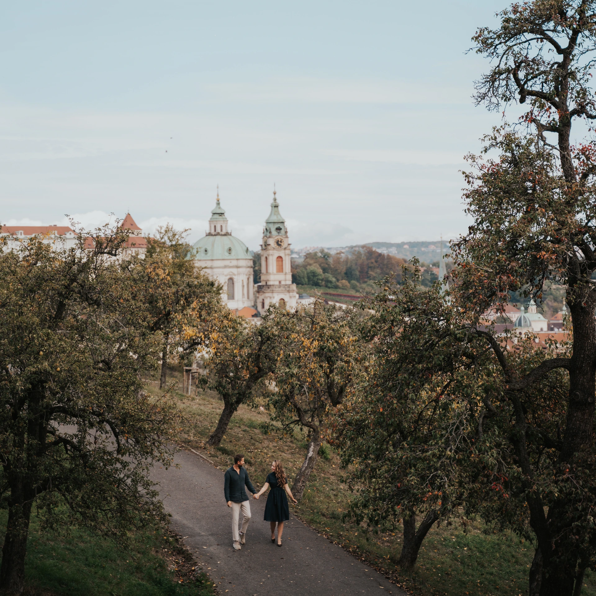 Couple walking hand in hand along a path between fruit trees, with a green church dome and tower above Prague’s red rooftops.