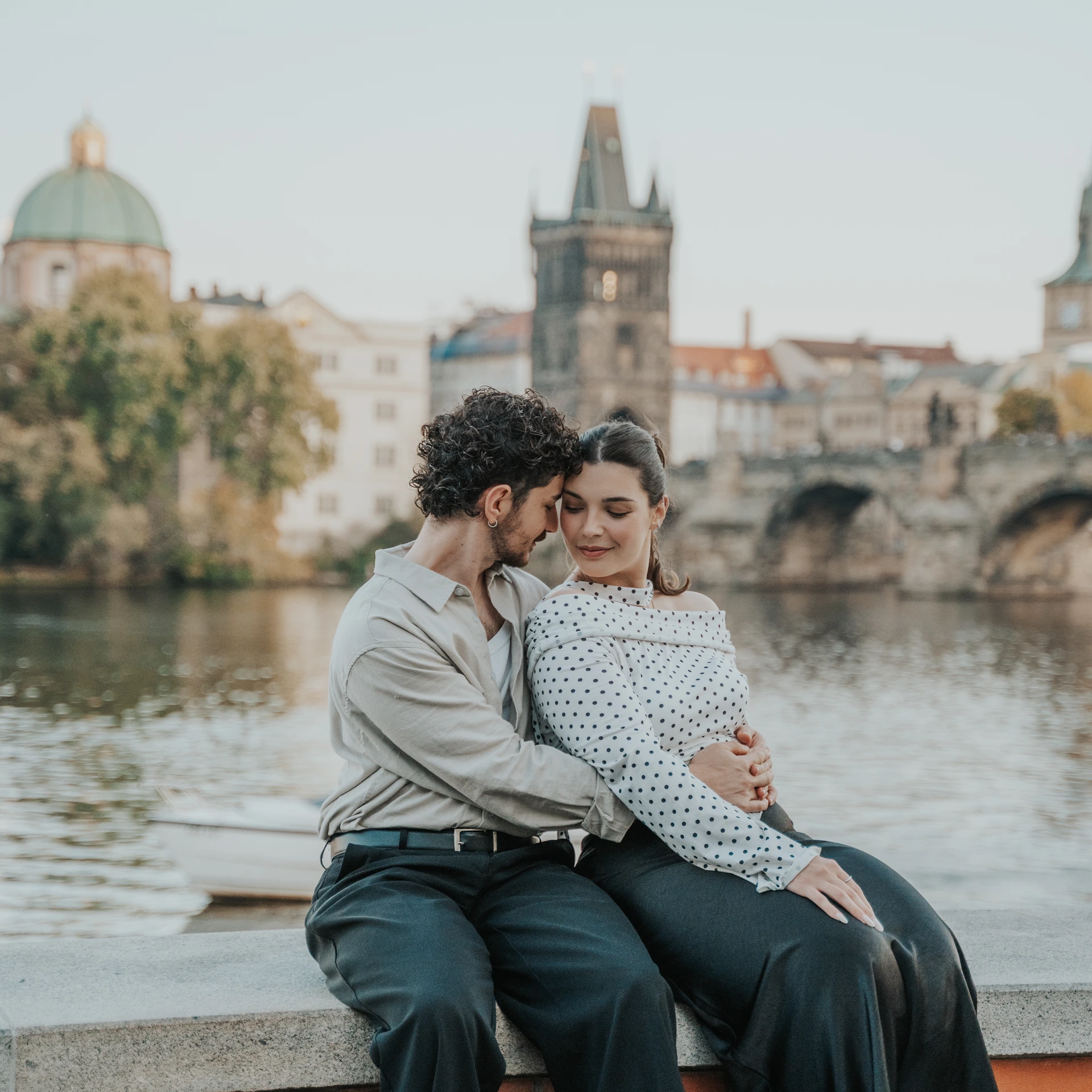Couple sitting close together beside the Vltava in Prague, with Charles Bridge and the Old Town in the background.