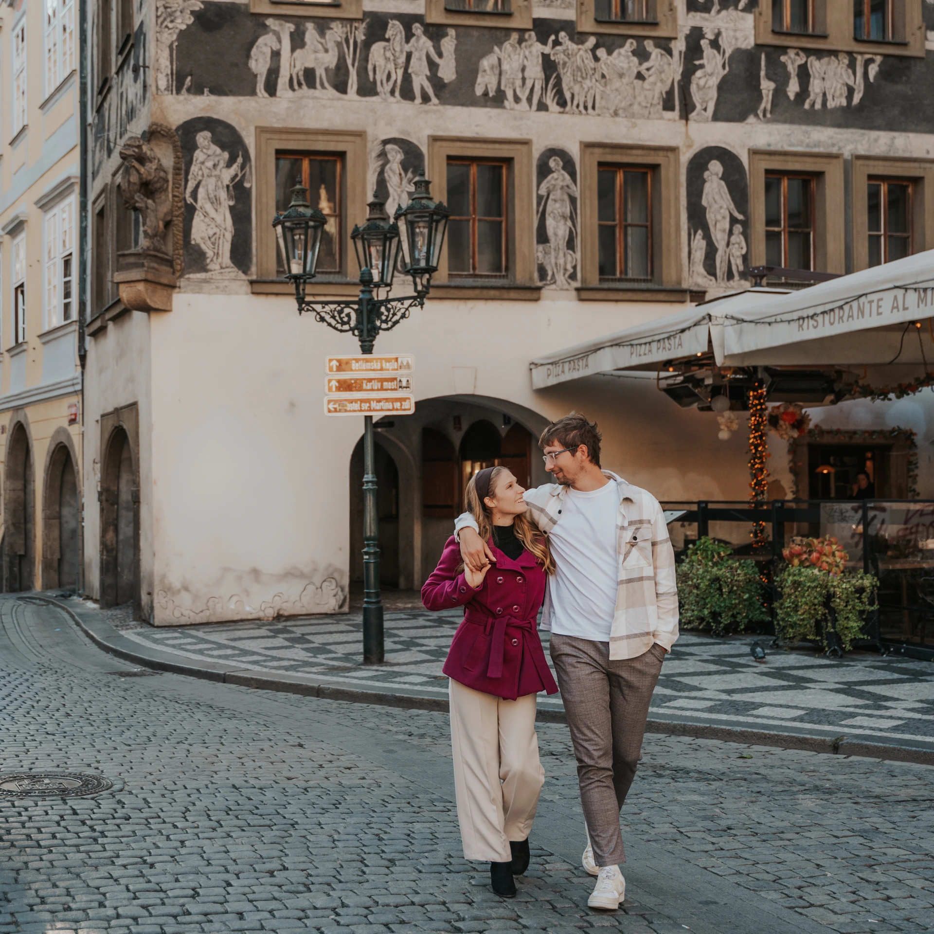 Couple walking arm in arm along a cobbled street in Prague Old Town, in front of a facade decorated with human figures.
