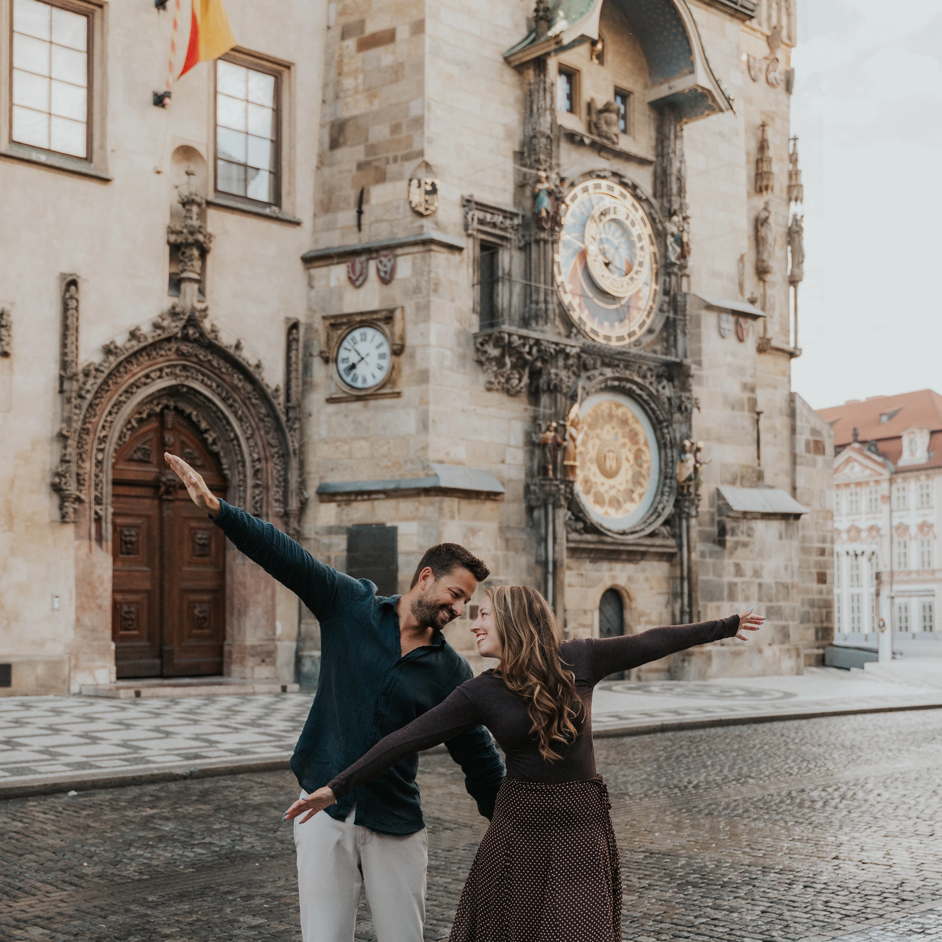 Couple dancing with outstretched arms in front of the Astronomical Clock in Old Town Square during a Prague photoshoot.