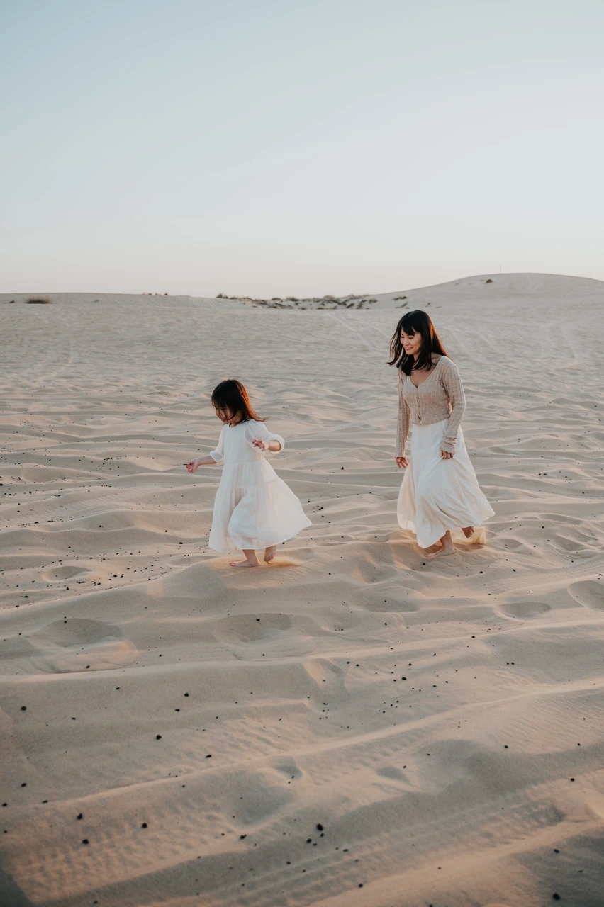Mothers day gift ideas Dubai - Mother and daughter in desert A mother and young daughter in white dresses walk barefoot together on soft Dubai desert sand at sunset.