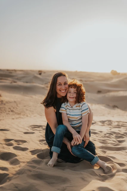 A mother and her curly-haired son sit close together on Dubai dunes in soft sunset light during a family session.