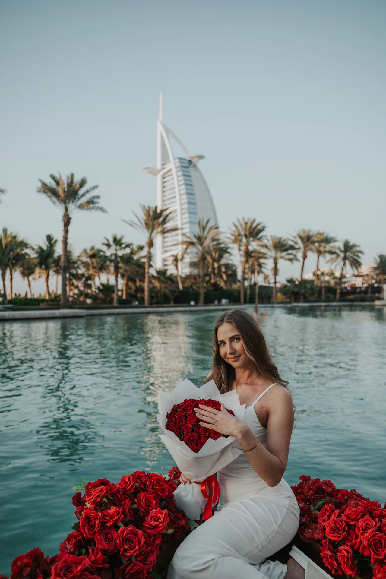 A woman holding a red rose bouquet sits on an abra in Souk Madinat with Burj Al Arab behind her.