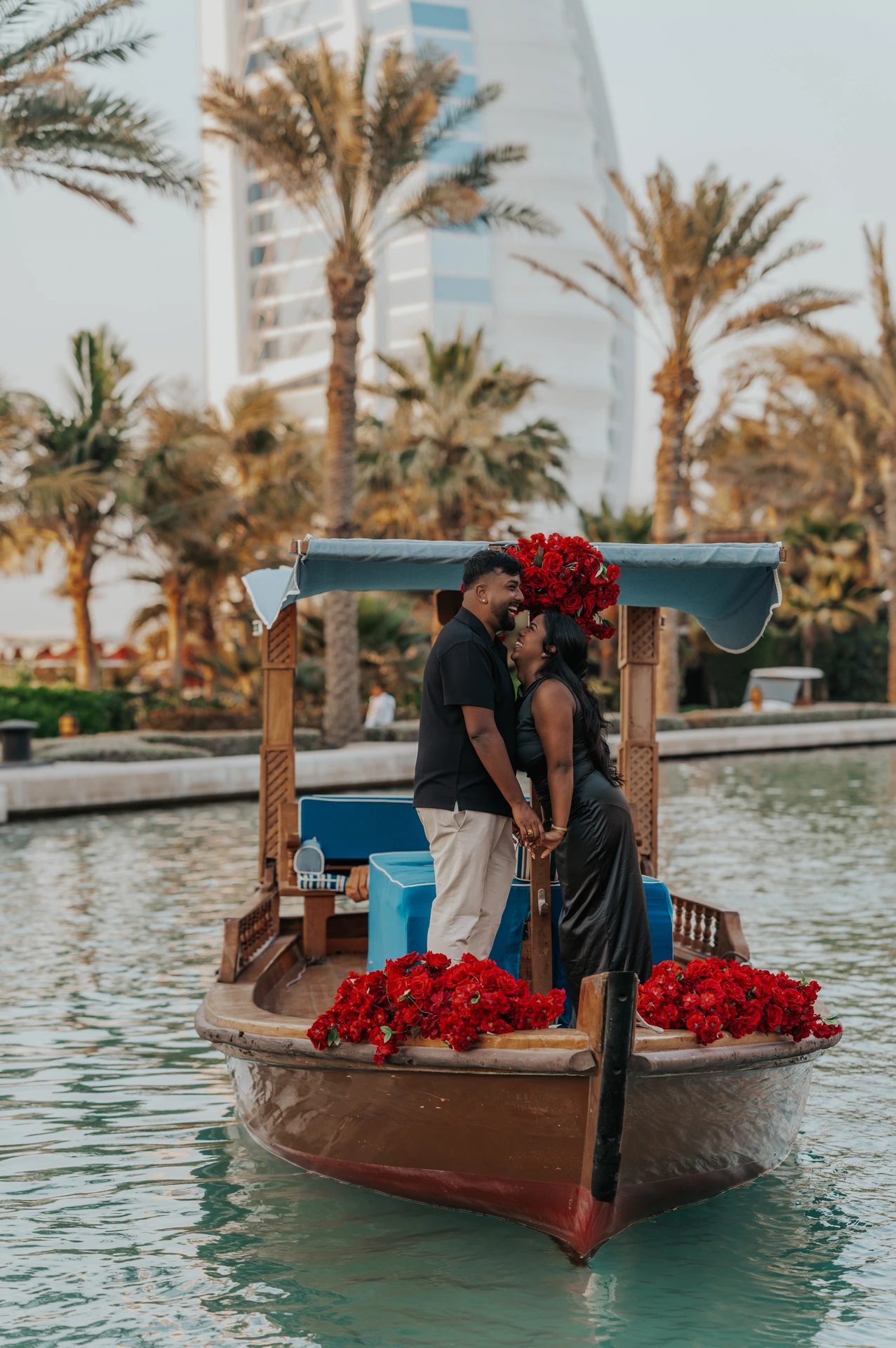 Abra Proposal at Souk Madinat Dubai - Joyful Couple on Abra A smiling couple in evening outfits stands on an abra decorated with red roses in Souk Madinat.