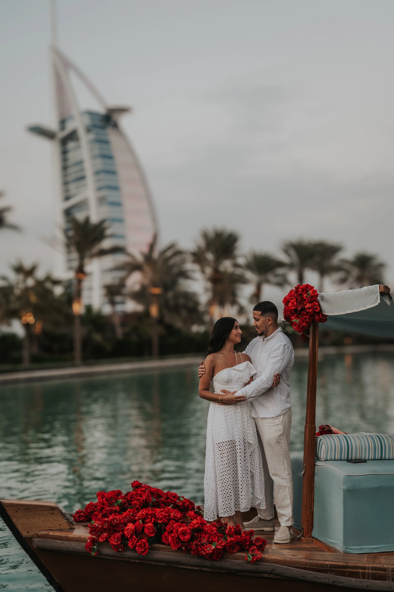 A couple stands together on an abra at Souk Madinat during sunset with floral decorations and calm water.