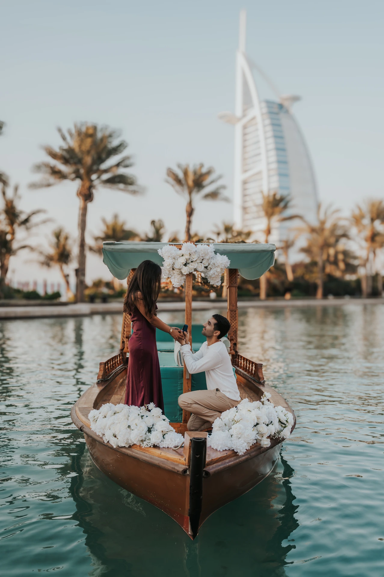 A groom-to-be kneels on a flower-decorated abra while proposing in Souk Madinat with Burj Al Arab behind.