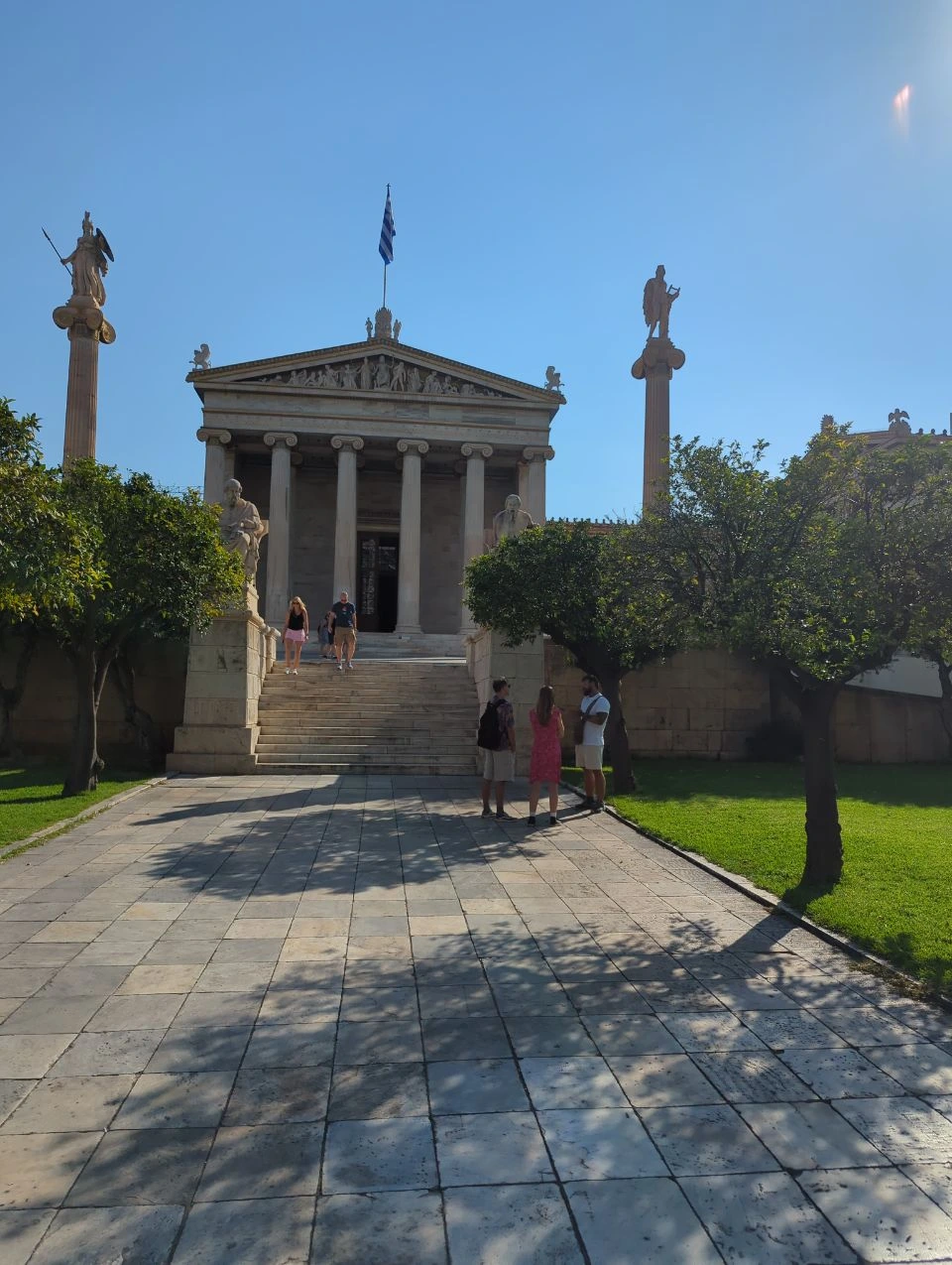 Athens Photo Tour - Academy Architecture Visitors explore the Academy of Athens, where statues, columns and a Greek flag frame the neoclassical facade.