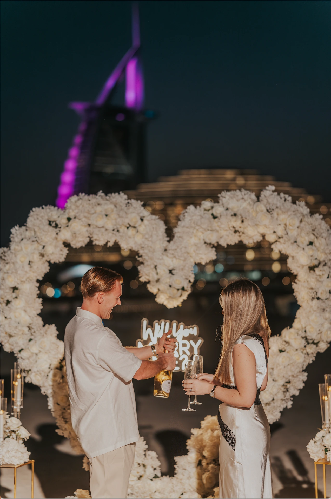 A couple celebrates with champagne beneath a white floral heart setup on Dubai beach at night, with Burj Al Arab lit in the background.