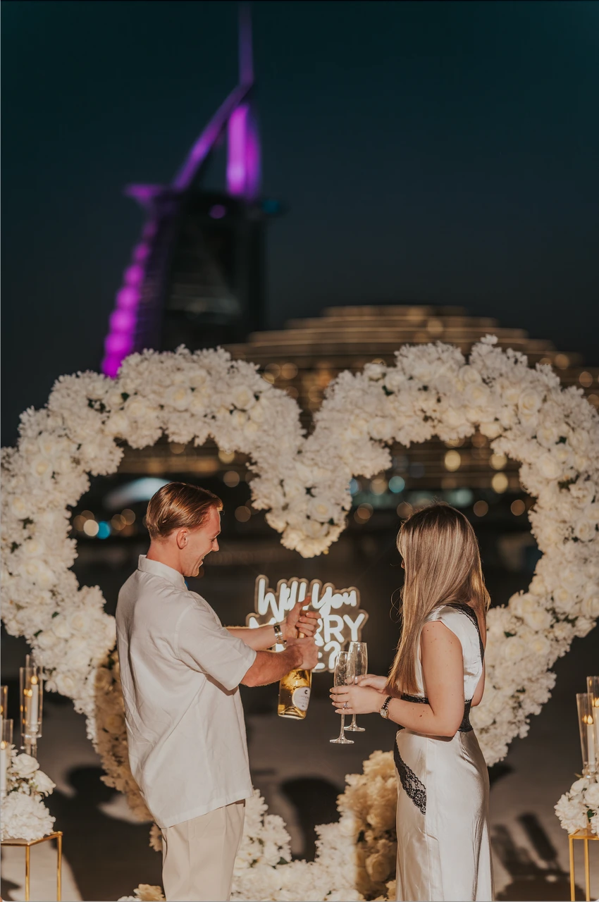 Heart Proposal Setup Dubai - champagne toast under white arch A couple celebrates with champagne beneath a white floral heart setup on Dubai beach at night, with Burj Al Arab lit in the background.