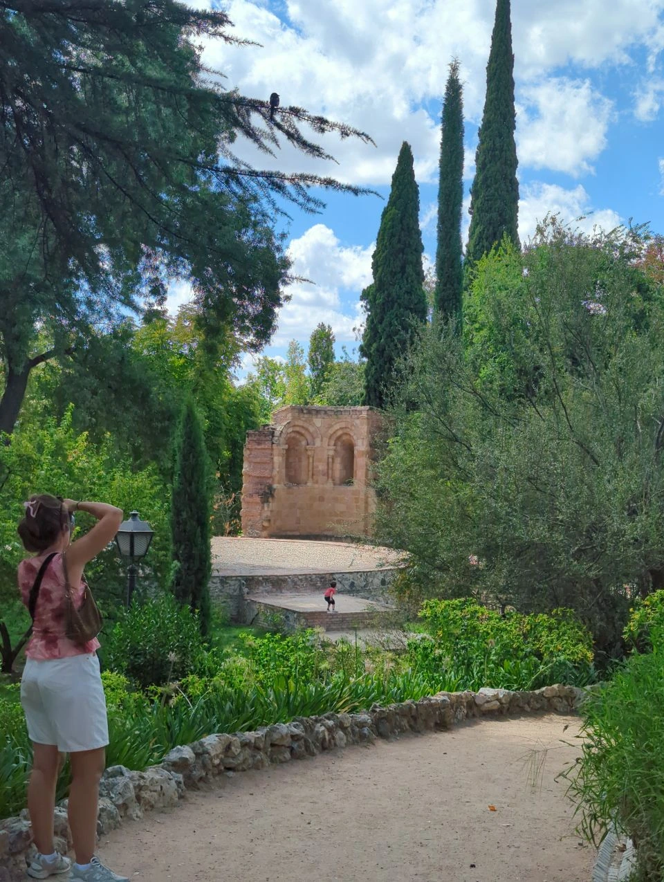 An adult photographs a child on a garden path beside greenery and old stonework at Montaña Artificial in Madrid.
