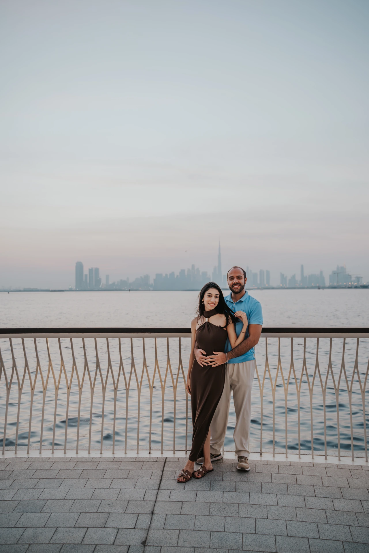 Affordable proposal Dubai - Skyline maternity-style couple portrait A couple poses by a Dubai waterfront railing with skyline towers behind them in soft evening haze.