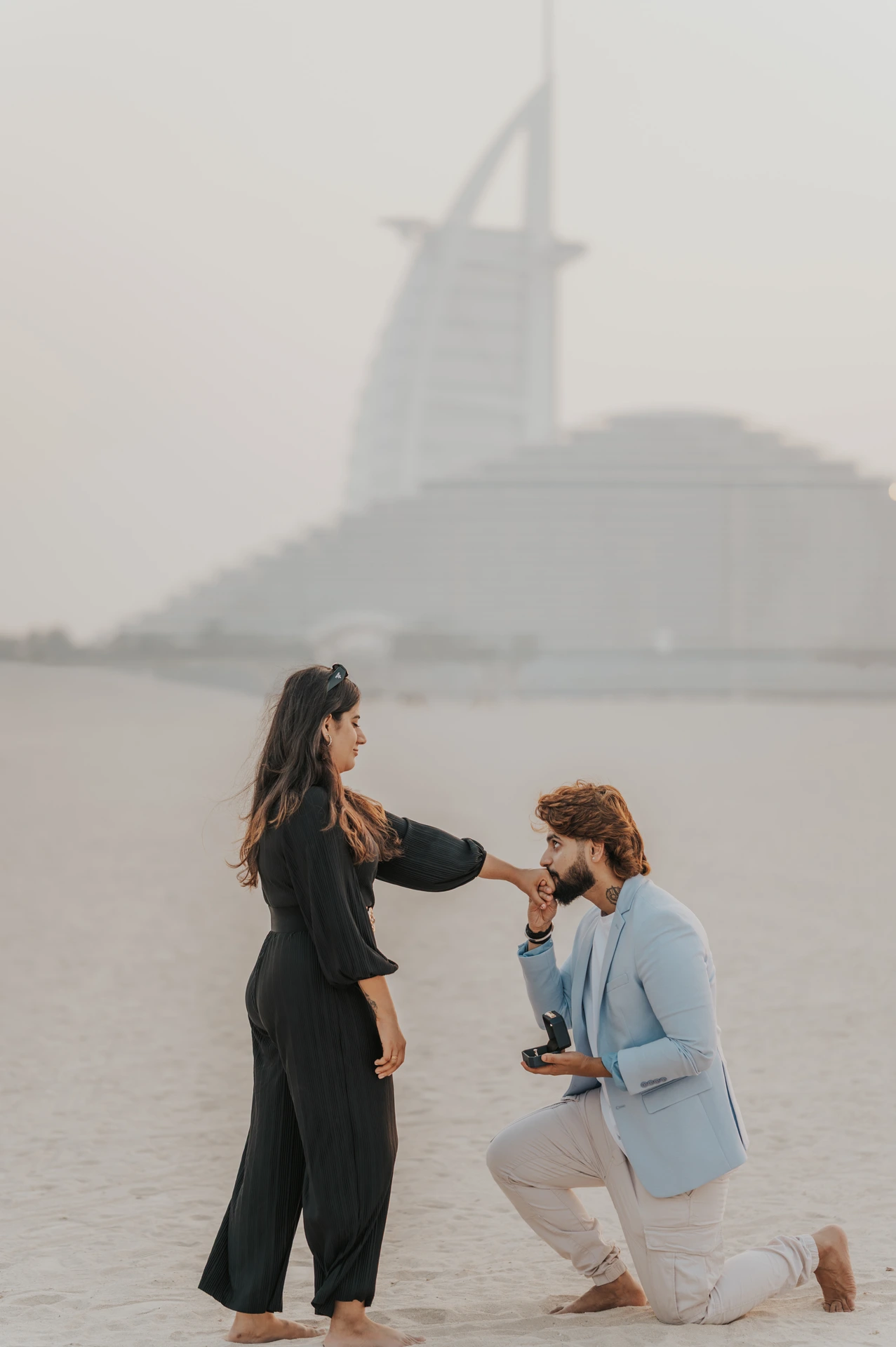A man kneels on the beach to propose while his partner smiles, with Burj Al Arab visible in the background.