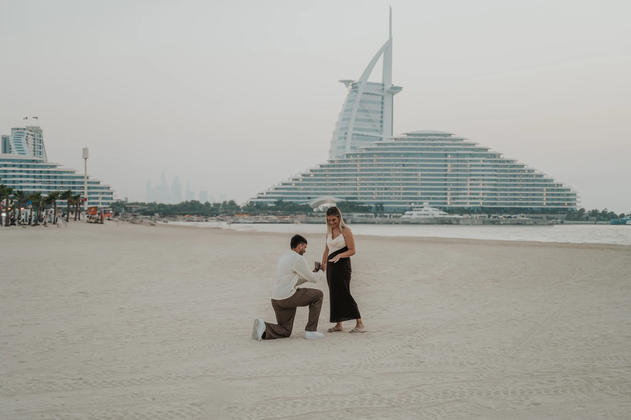 Proposal at Shimmers - wide beach proposal frame A man proposes on the sand near Shimmers in Dubai while his partner stands smiling, captured in a wide beach composition.