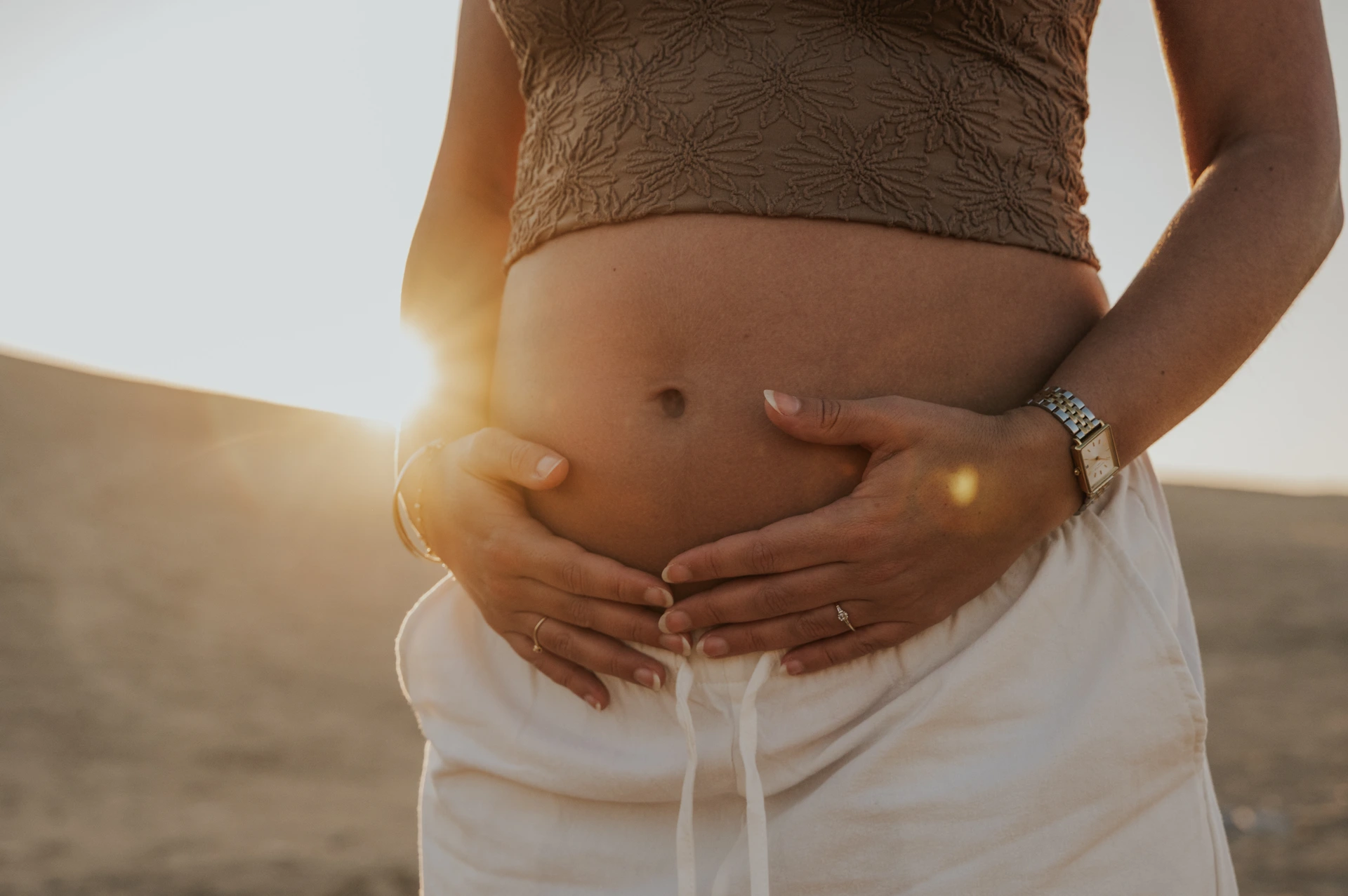 Close-up eines schwangeren Bauches während eines Babybauchshootings bei Sonnenuntergang in der Wüste von Dubai, mit der Sonne, die hinter den Dünen auf ihren Bauch scheint.