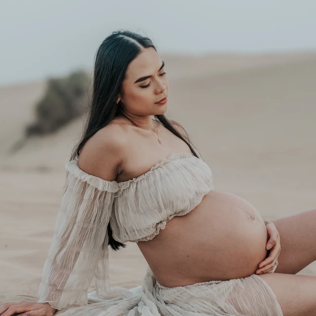 Babybauchshooting Dubai Schwangere Frau, die während eines Babybauchshootings in der Wüste von Dubai im Sand sitzt.
