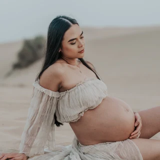 Schwangere Frau, die während eines Babybauchshootings in der Wüste von Dubai im Sand sitzt.