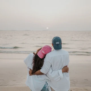 Schwangeres Paar mit „Mom“- und „Dad“-Kappen während eines Babybauchshootings am Strand von Dubai bei Sonnenuntergang.
