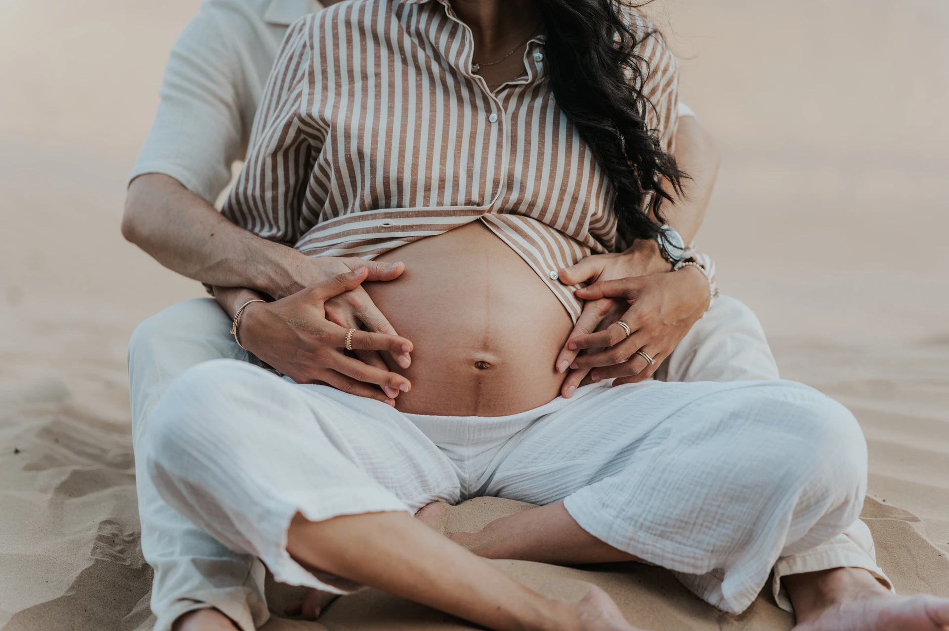 Paar, das während eines Babybauchshootings in der Wüste von Dubai im Sand sitzt und gemeinsam den schwangeren Bauch hält.
