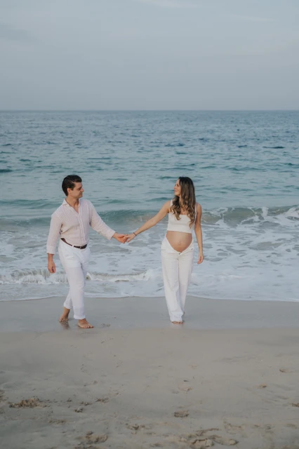 Couple in white outfits holding hands on a beach shoreline in Dubai during a relaxed maternity photoshoot.