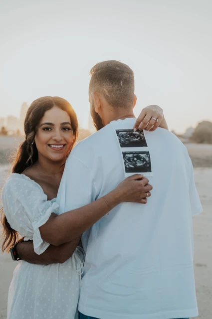 Smiling mother-to-be hugging her partner while showing ultrasound prints on a Dubai beach, for the Babymoon in Dubai guide.